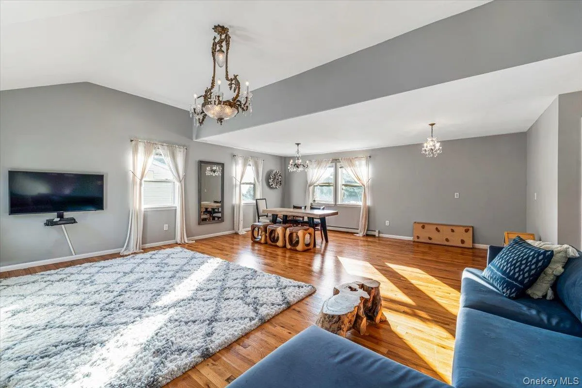 Spacious open-concept room featuring vaulted ceilings, wood-finish flooring, multiple windows with white curtains, and a decorative chandelier Spacious open-concept room featuring vaulted ceilings, wood-finish flooring, multiple windows with white curtains, and a decorative chandelier