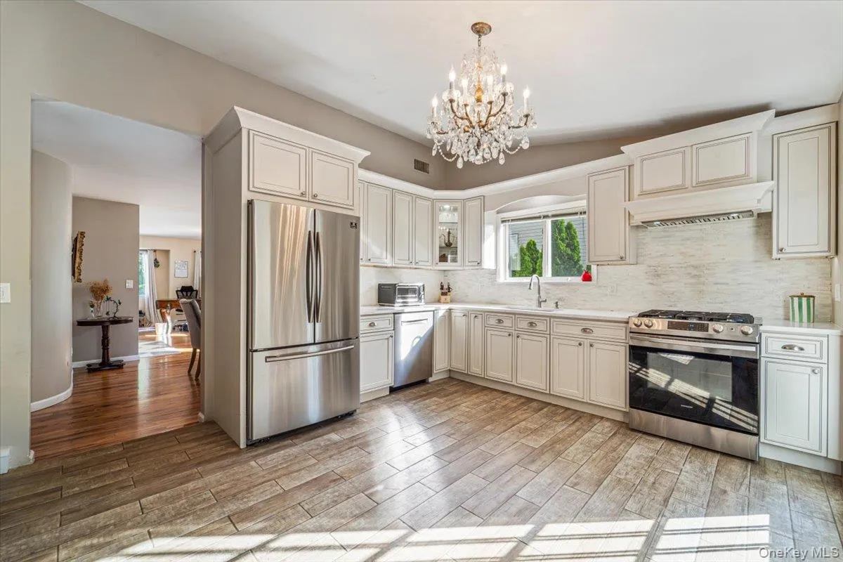 Kitchen featuring white cabinetry, stainless steel appliances, a crystal chandelier, wood-finish flooring, and a white tile backsplash Kitchen featuring white cabinetry, stainless steel appliances, a crystal chandelier, wood-finish flooring, and a white tile backsplash