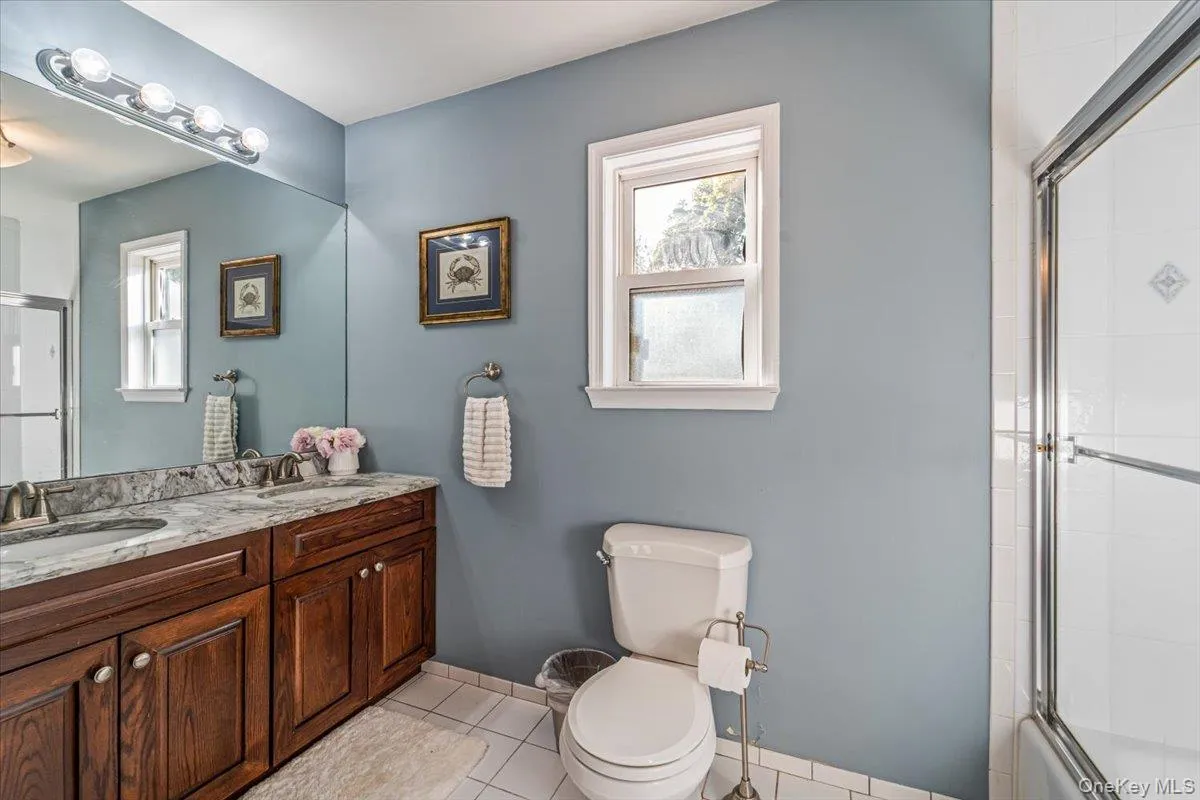 Bathroom featuring a dual-sink vanity with a stone countertop and wood cabinetry, a single window, and a shower with a sliding glass door Bathroom featuring a dual-sink vanity with a stone countertop and wood cabinetry, a single window, and a shower with a sliding glass door