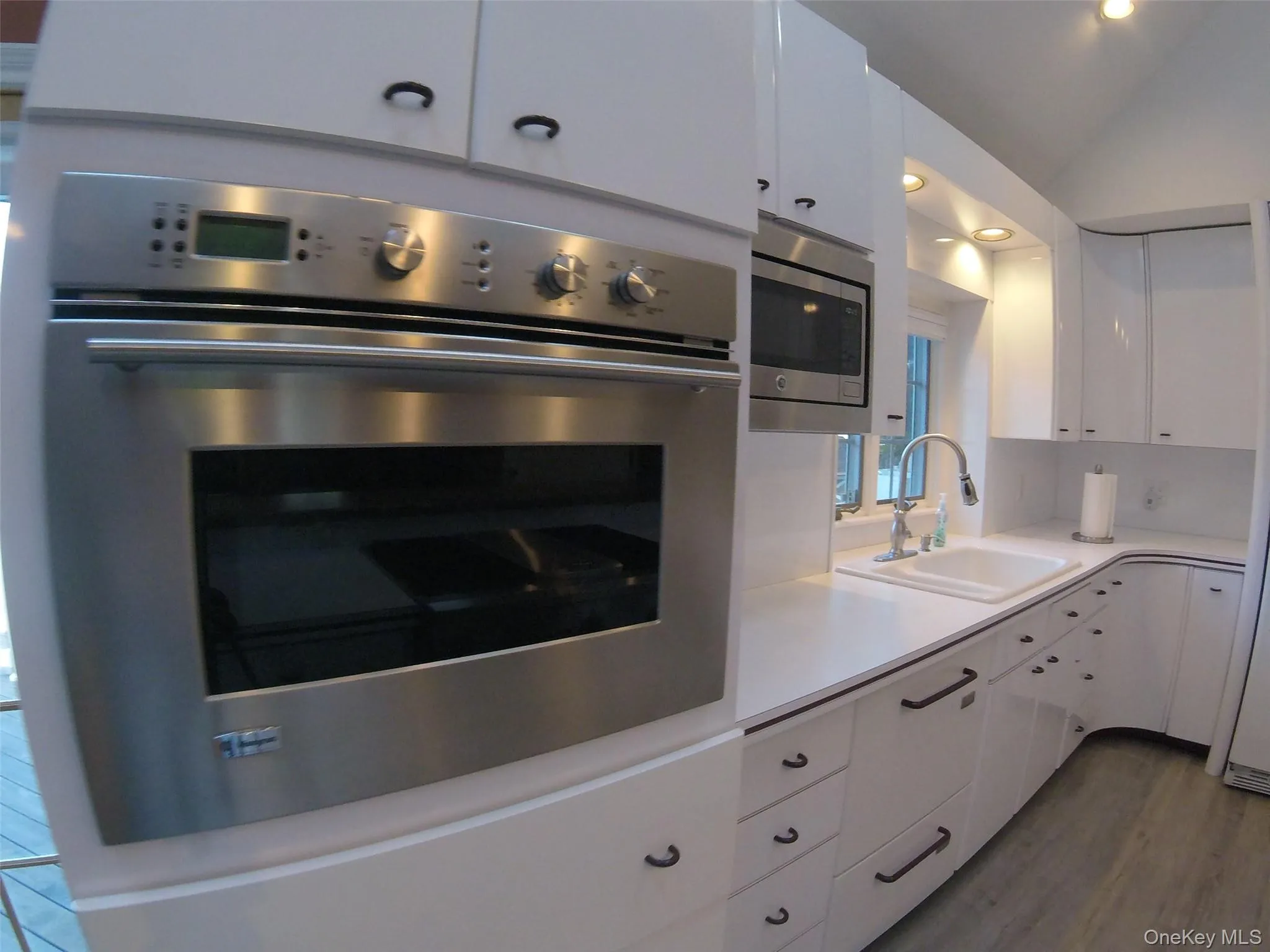 Built-in stainless steel oven and microwave, white cabinetry with black hardware, white solid surface countertops, a white sink with a gooseneck faucet, and wood-finish flooring Built-in stainless steel oven and microwave, white cabinetry with black hardware, white solid surface countertops, a white sink with a gooseneck faucet, and wood-finish flooring