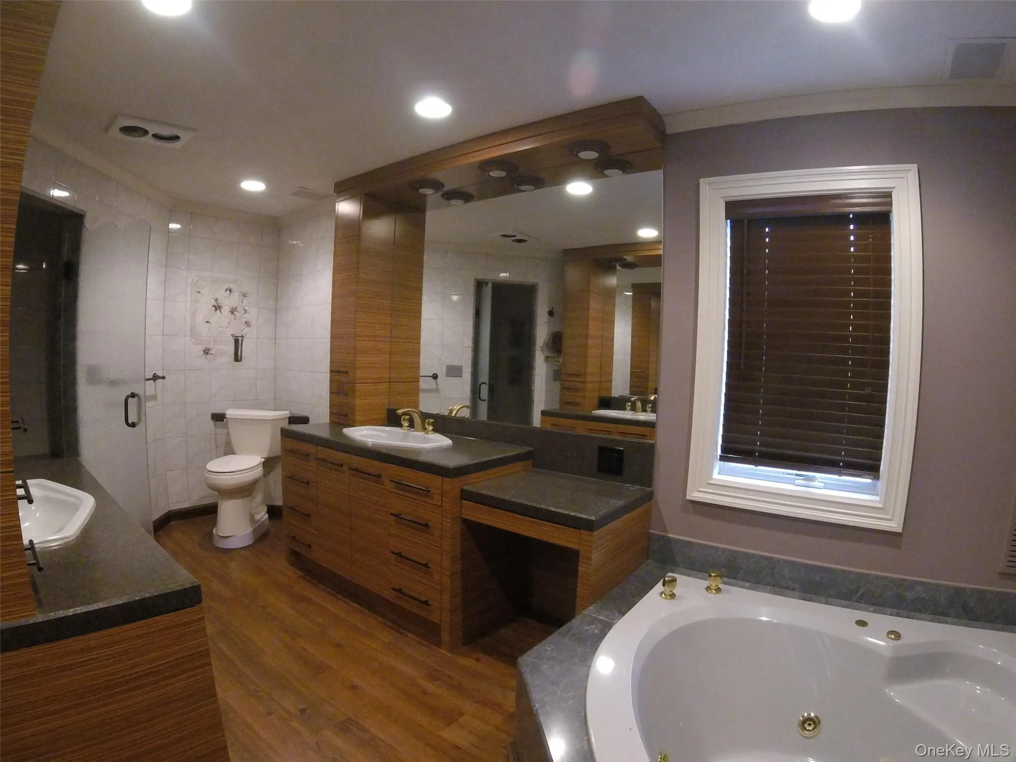 Spacious bathroom featuring a built-in jetted tub, wood-finish vanity with integrated sink, and a large mirror with integrated lighting Spacious bathroom featuring a built-in jetted tub, wood-finish vanity with integrated sink, and a large mirror with integrated lighting