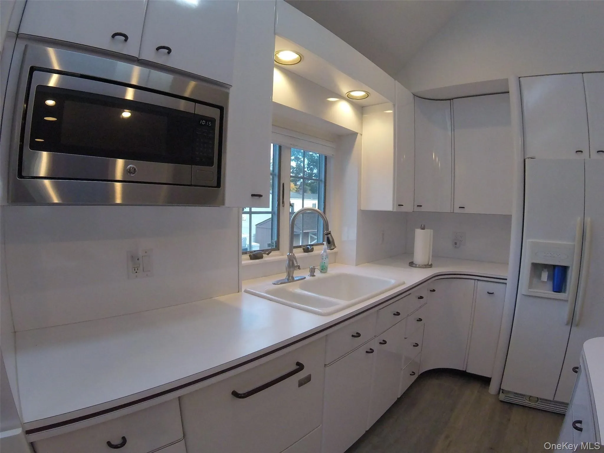 Bright kitchen featuring white cabinetry, a double basin sink with a gooseneck faucet, an over-range stainless steel microwave, solid surface countertops, and recessed lighting Bright kitchen featuring white cabinetry, a double basin sink with a gooseneck faucet, an over-range stainless steel microwave, solid surface countertops, and recessed lighting