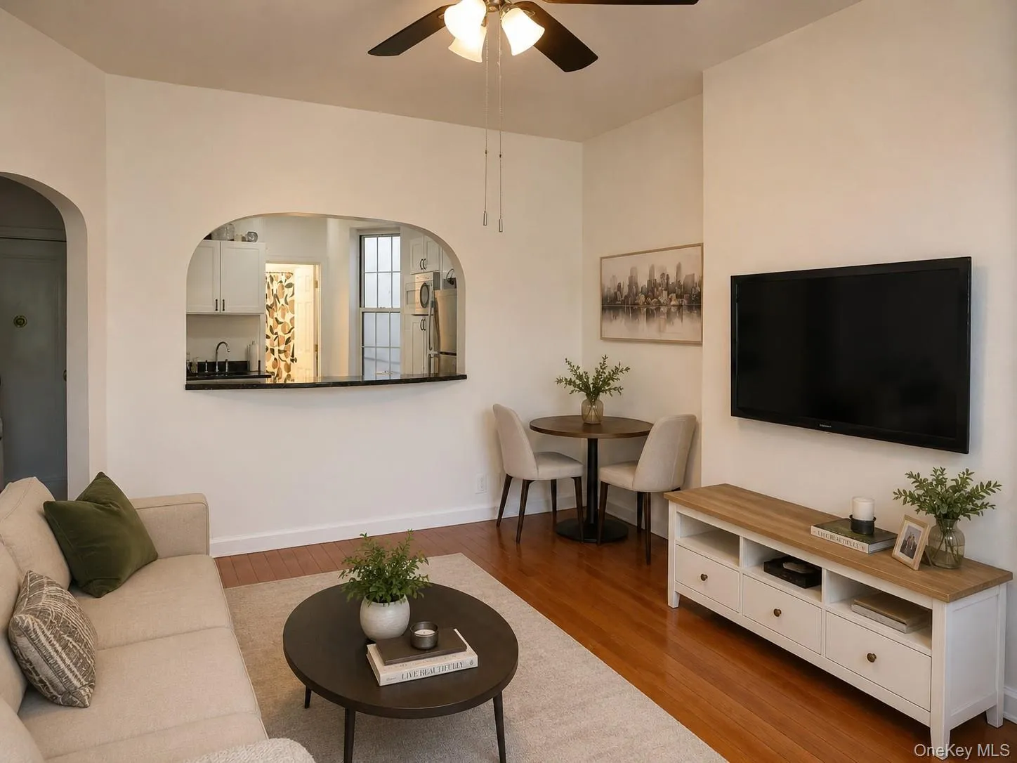 Open concept living area featuring wood-finish flooring, a ceiling fan with integrated lighting, and an arched pass-through window to the kitchen. (virtually staged) Open concept living area featuring wood-finish flooring, a ceiling fan with integrated lighting, and an arched pass-through window to the kitchen. (virtually staged)