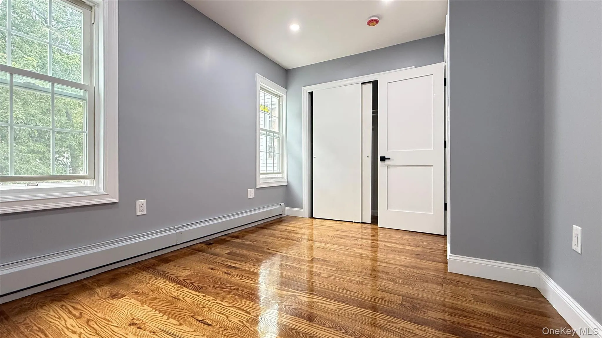 Spacious room featuring wood-finish flooring, two large windows, modern baseboard heating, and white trim Spacious room featuring wood-finish flooring, two large windows, modern baseboard heating, and white trim