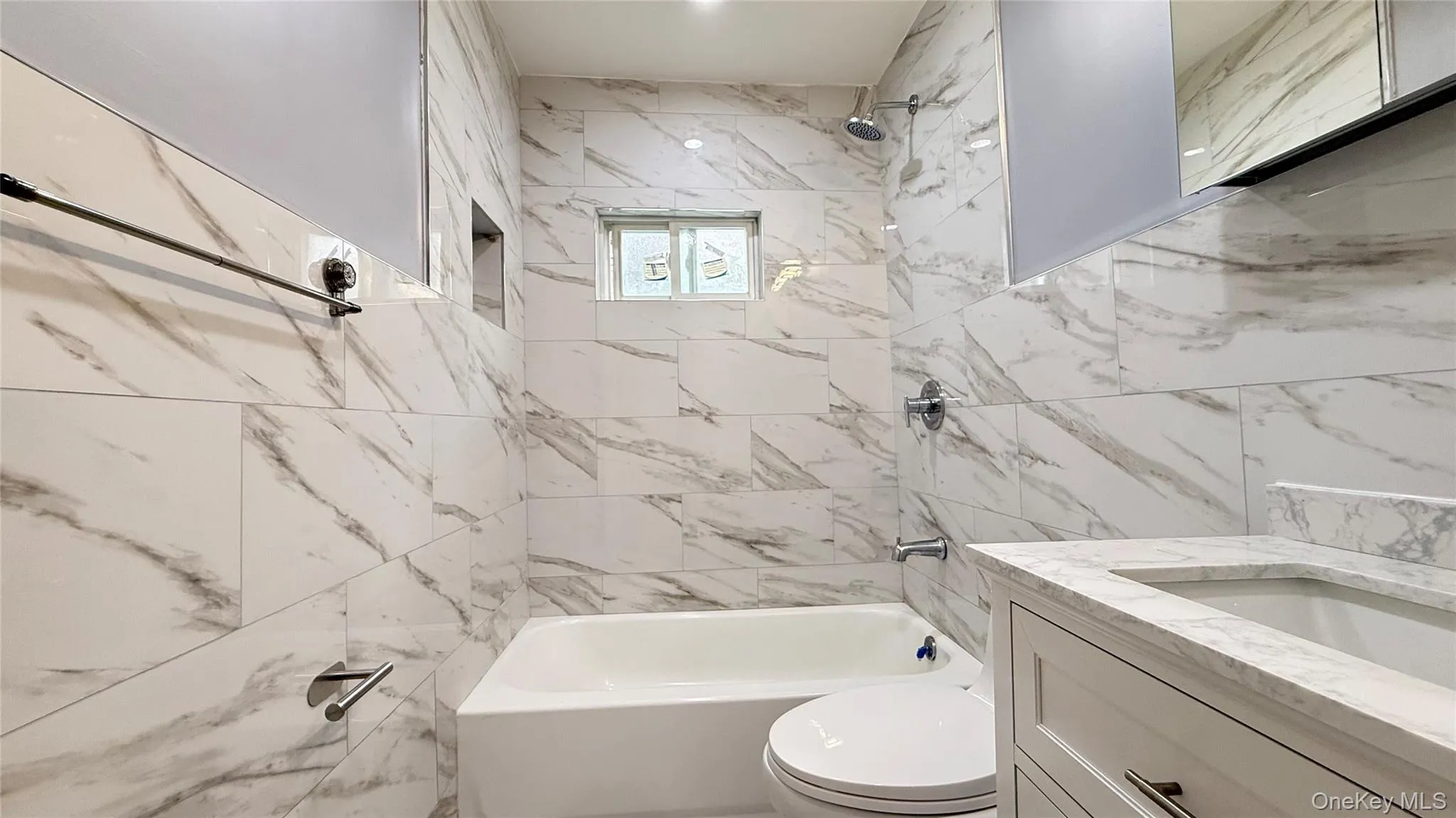 Bathroom featuring large format marbled wall tiling, a white built-in bathtub, a window with obscure glass, a recessed shower niche, and a white vanity with an integrated sink Bathroom featuring large format marbled wall tiling, a white built-in bathtub, a window with obscure glass, a recessed shower niche, and a white vanity with an integrated sink