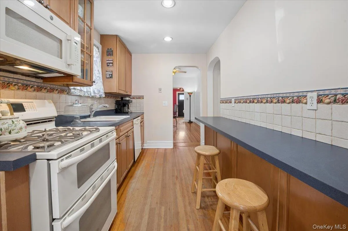 Galley kitchen featuring wood-finish flooring, light wood cabinetry, white tile backsplash with decorative border, and a built-in double oven range Galley kitchen featuring wood-finish flooring, light wood cabinetry, white tile backsplash with decorative border, and a built-in double oven range