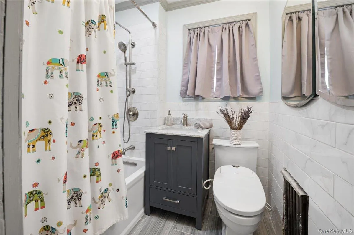 Bathroom featuring a tub/shower combination with a stainless steel shower fixture, a dark gray vanity with a light countertop, a toilet, and light-toned wall tiling Bathroom featuring a tub/shower combination with a stainless steel shower fixture, a dark gray vanity with a light countertop, a toilet, and light-toned wall tiling