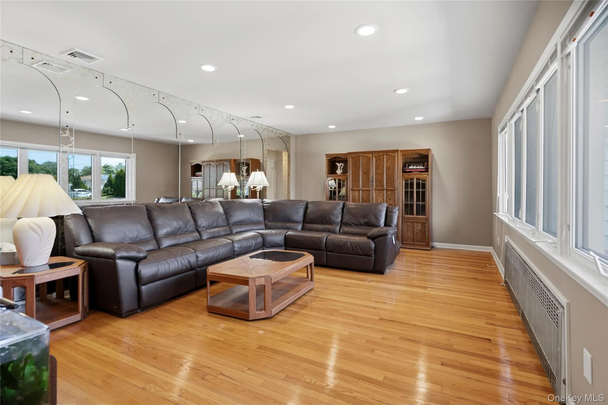 Spacious living area featuring polished wood flooring, recessed lighting, a large built-in mirror installation with arched accents, and multiple windows providing natural illumination Spacious living area featuring polished wood flooring, recessed lighting, a large built-in mirror installation with arched accents, and multiple windows providing natural illumination