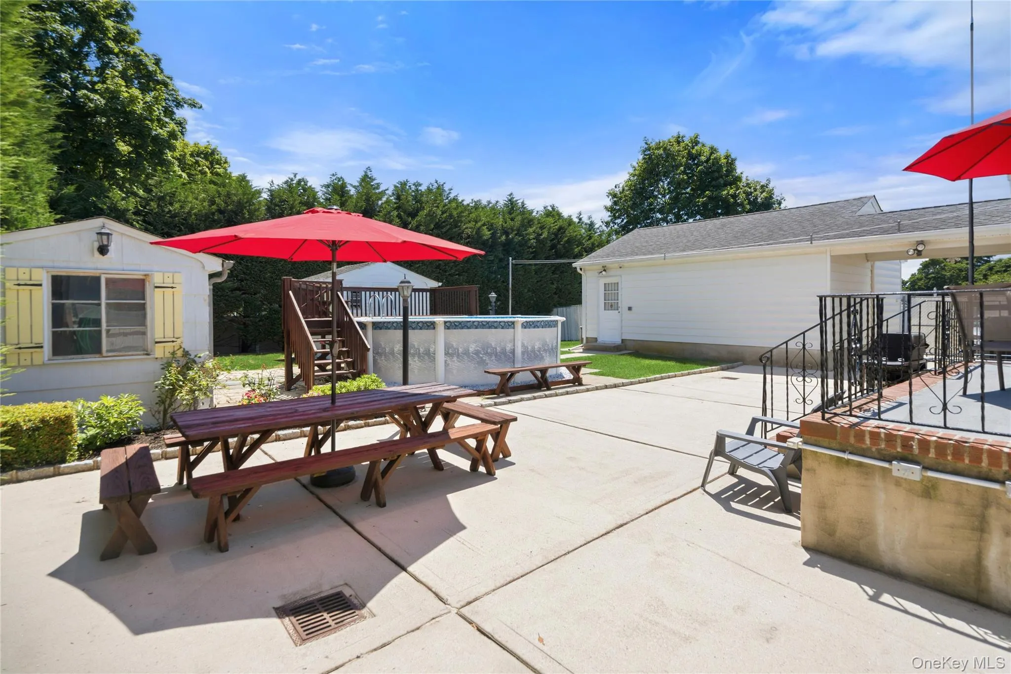 Spacious concrete patio featuring an above-ground pool, a white ancillary structure with yellow shutters, a separate white garage structure, and a raised patio area with wrought iron railings Spacious concrete patio featuring an above-ground pool, a white ancillary structure with yellow shutters, a separate white garage structure, and a raised patio area with wrought iron railings