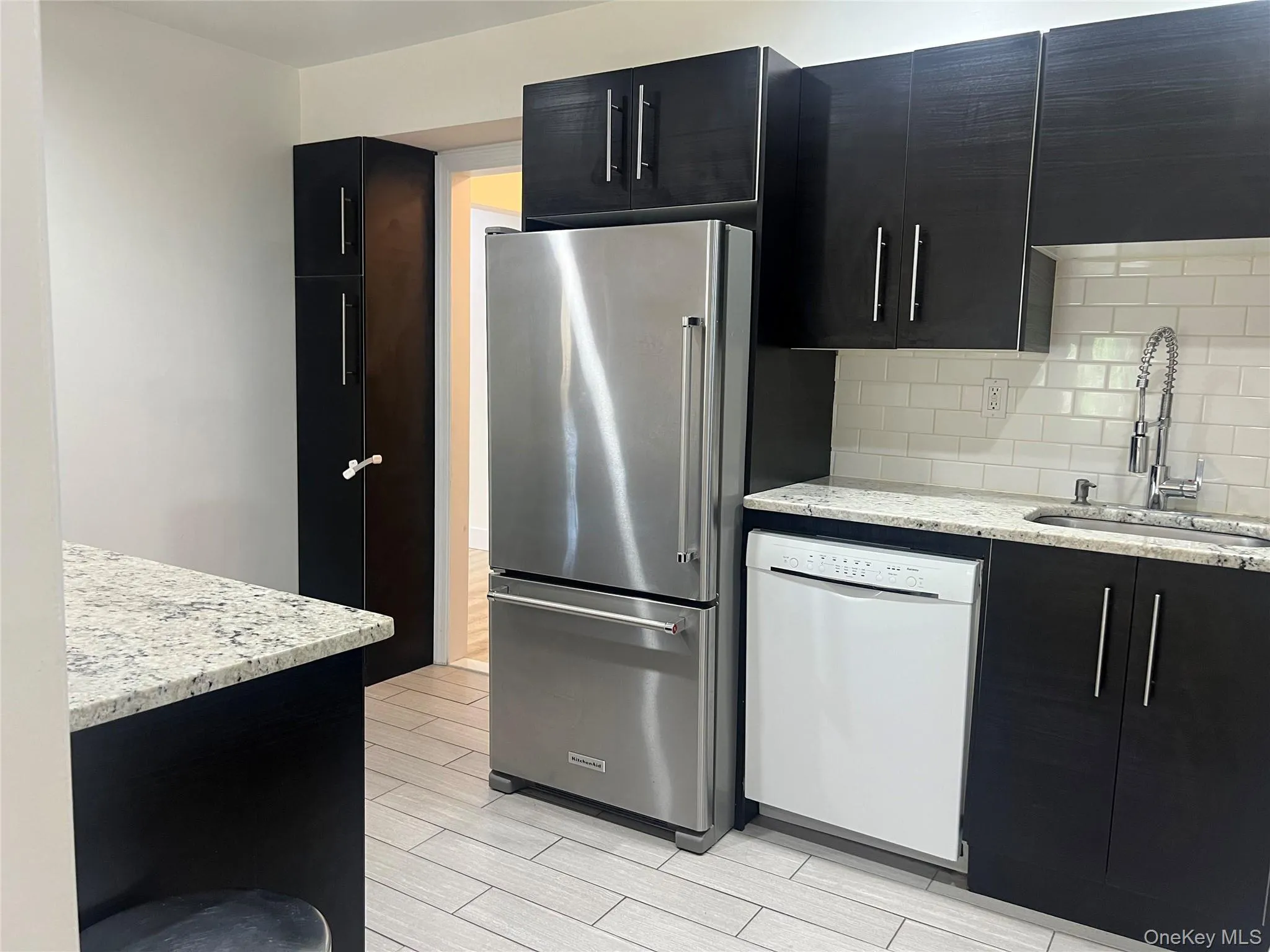 Modern kitchen featuring dark cabinetry, stainless steel refrigerator, white subway tile backsplash, light granite countertops, and light tile flooring Modern kitchen featuring dark cabinetry, stainless steel refrigerator, white subway tile backsplash, light granite countertops, and light tile flooring