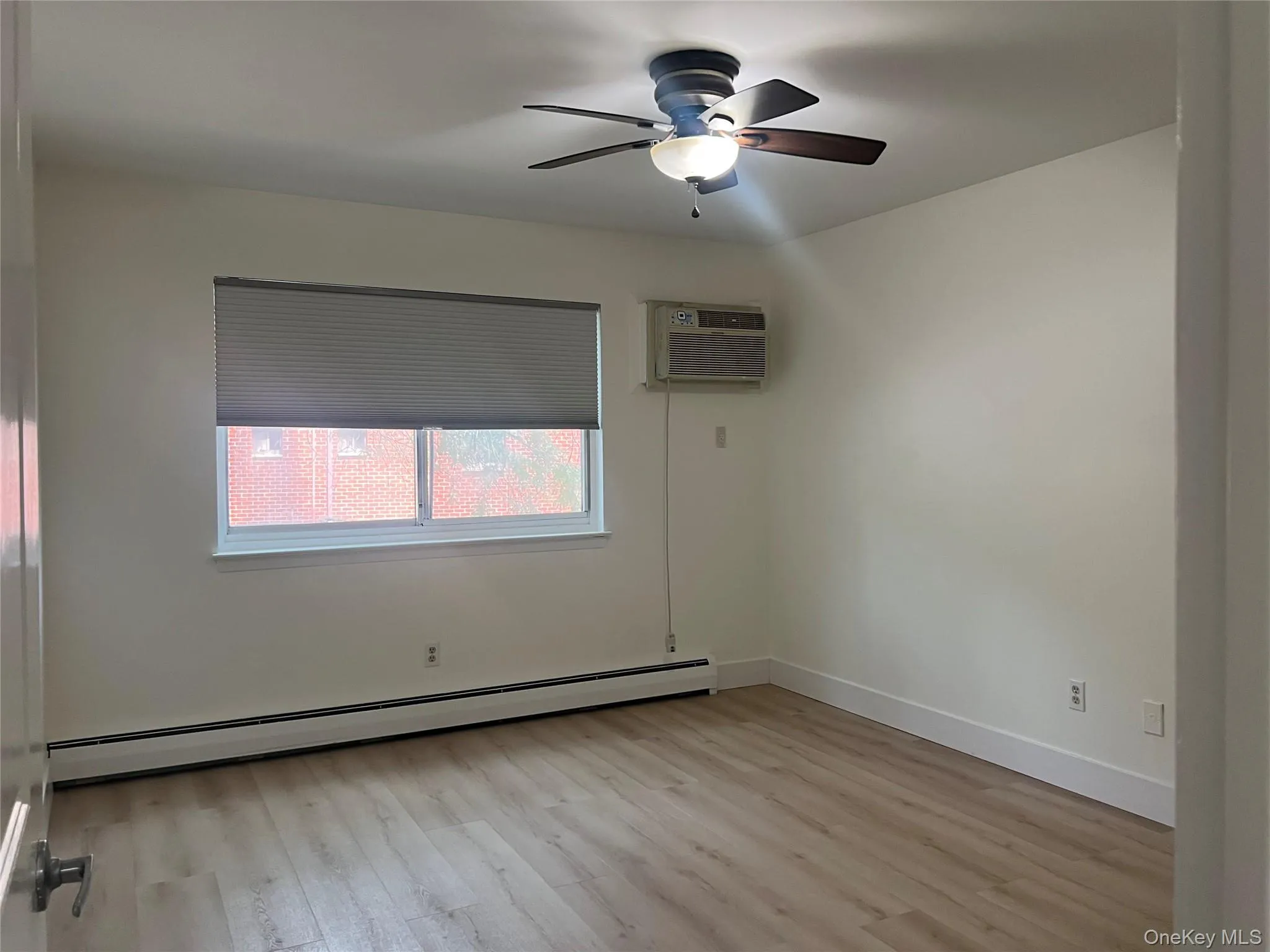 Room featuring wood-finish flooring, a ceiling fan with integrated lighting, window with cellular blinds, baseboard heating, and an installed air conditioning unit Room featuring wood-finish flooring, a ceiling fan with integrated lighting, window with cellular blinds, baseboard heating, and an installed air conditioning unit