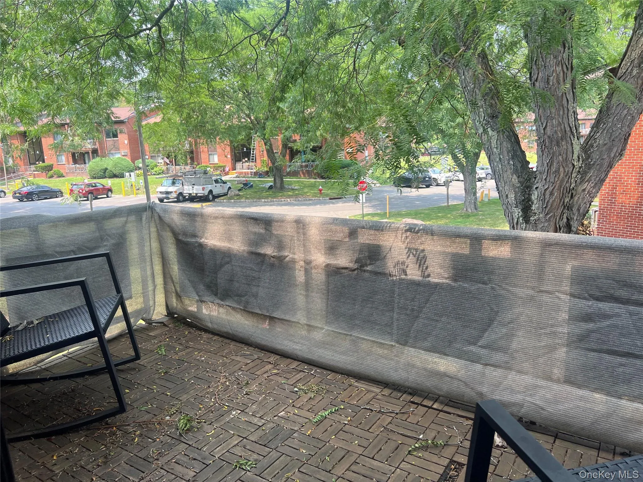 Exterior balcony with paver-style flooring, partial privacy screening, mature trees, and a brick-faced building exterior Exterior balcony with paver-style flooring, partial privacy screening, mature trees, and a brick-faced building exterior