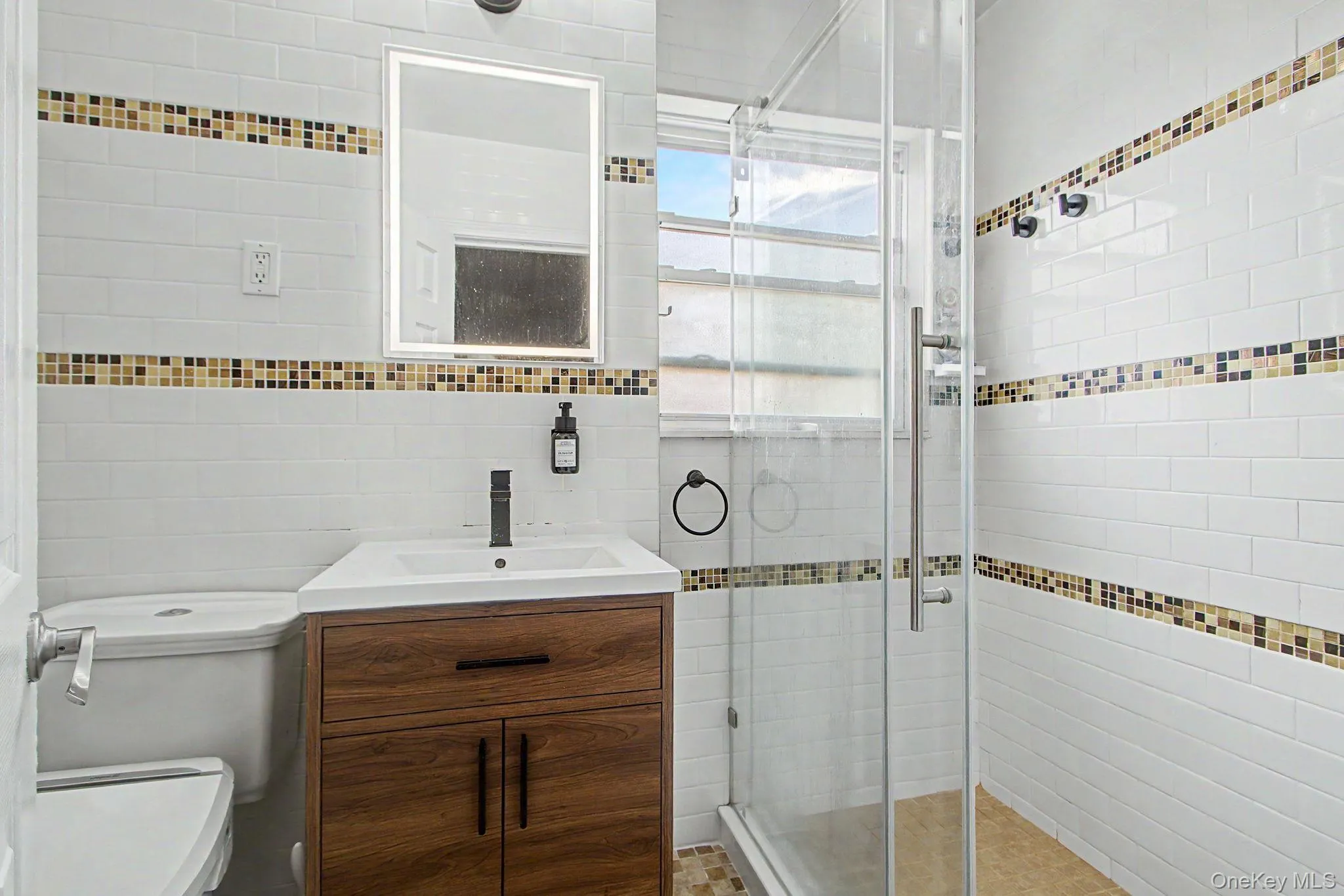 Bathroom featuring a wood-finish vanity with an integrated sink, a frameless shower enclosure, and white subway tile walls with decorative mosaic accents Bathroom featuring a wood-finish vanity with an integrated sink, a frameless shower enclosure, and white subway tile walls with decorative mosaic accents