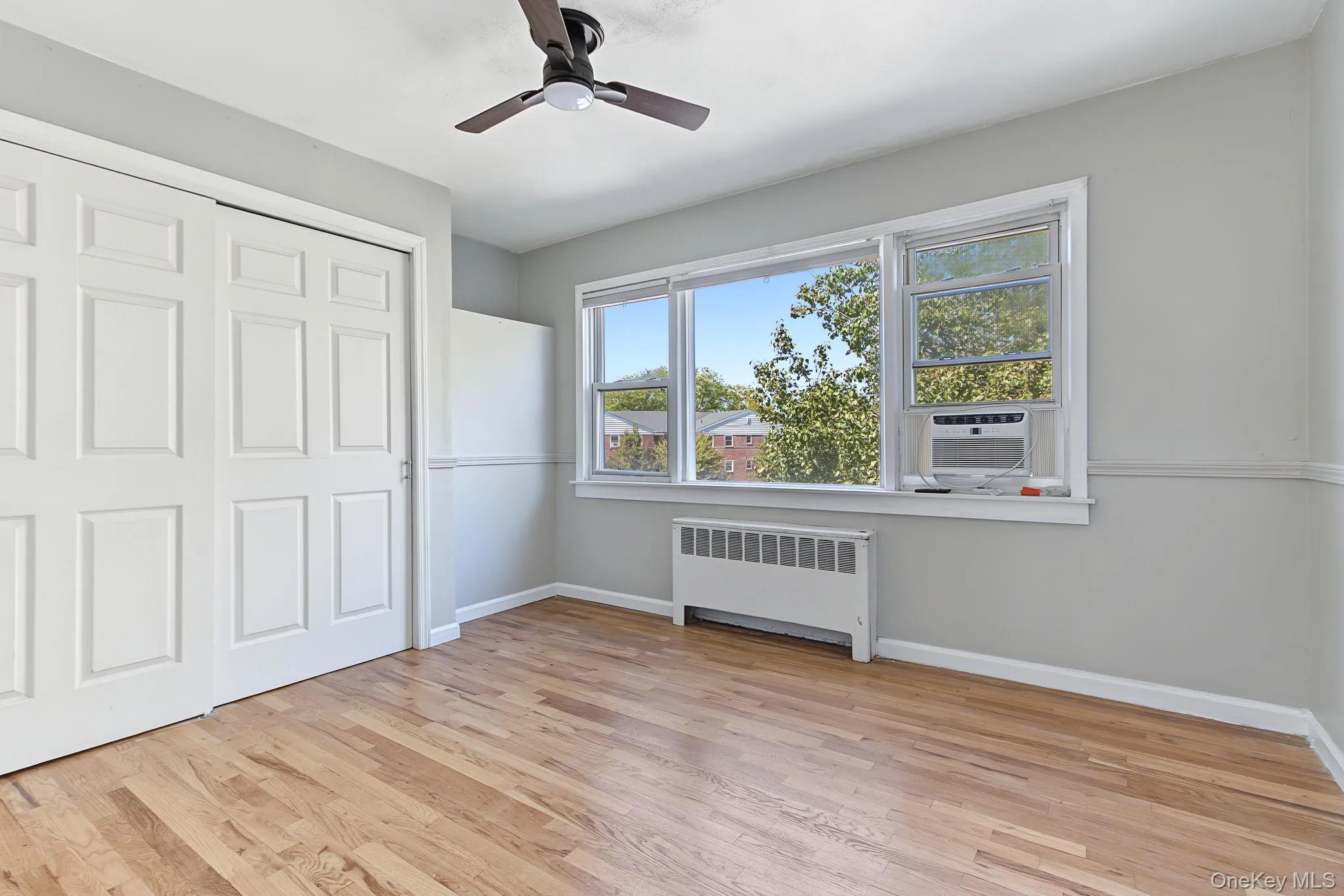 Hardwood flooring, multi-panel bi-fold closet doors, ceiling fan with integrated lighting, window-mounted air conditioning unit, and a window with a radiator below Hardwood flooring, multi-panel bi-fold closet doors, ceiling fan with integrated lighting, window-mounted air conditioning unit, and a window with a radiator below