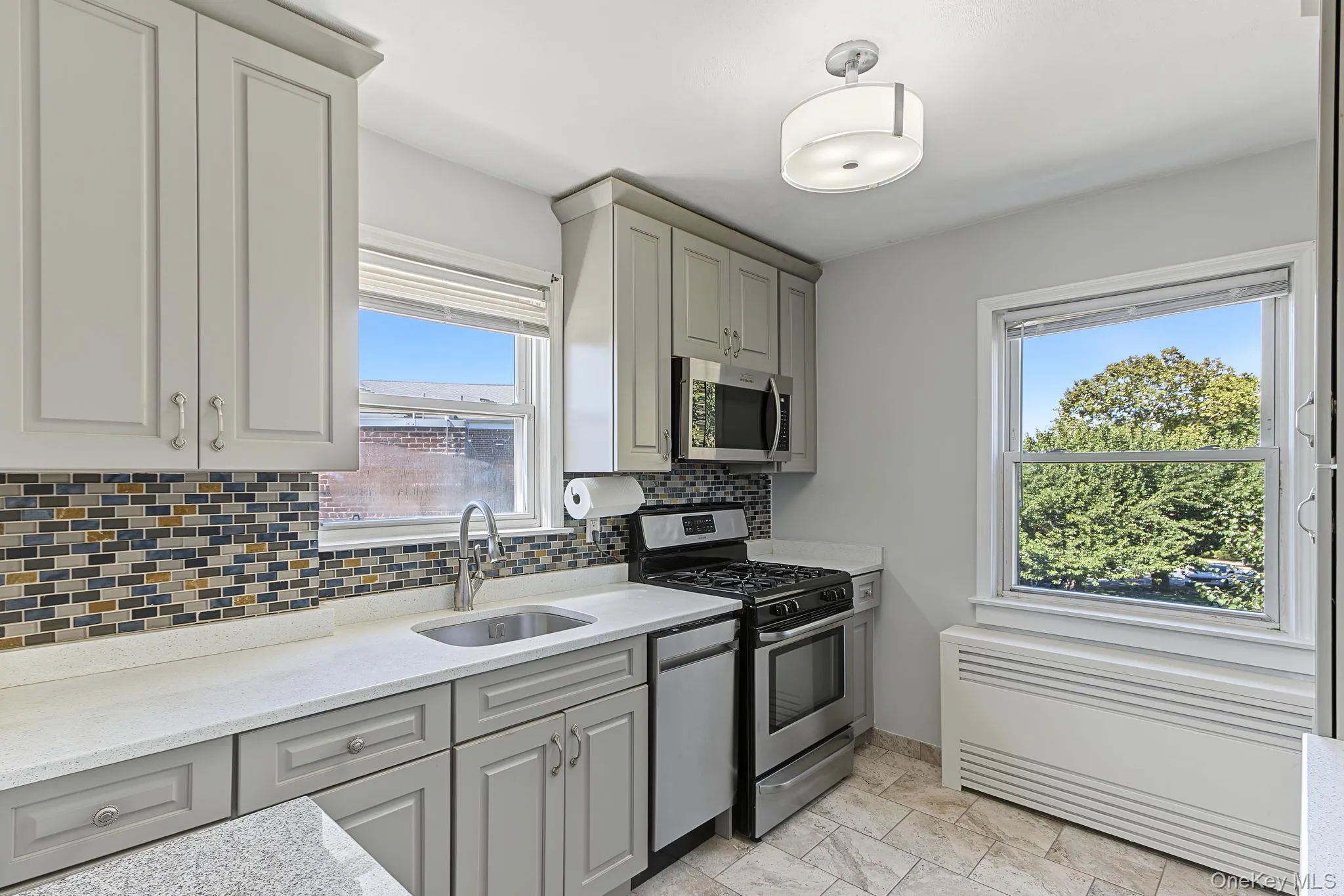 Light gray cabinetry with crown molding, stainless steel appliances, mosaic tile backsplash, white countertops, and tiled flooring Light gray cabinetry with crown molding, stainless steel appliances, mosaic tile backsplash, white countertops, and tiled flooring