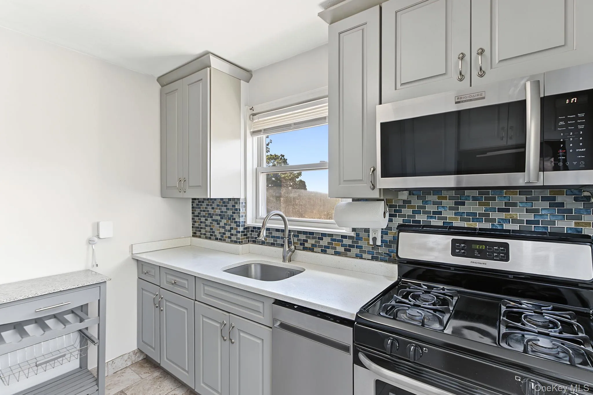 White countertops, stainless steel appliances, mosaic tile backsplash, gray cabinetry, and a stainless steel sink White countertops, stainless steel appliances, mosaic tile backsplash, gray cabinetry, and a stainless steel sink