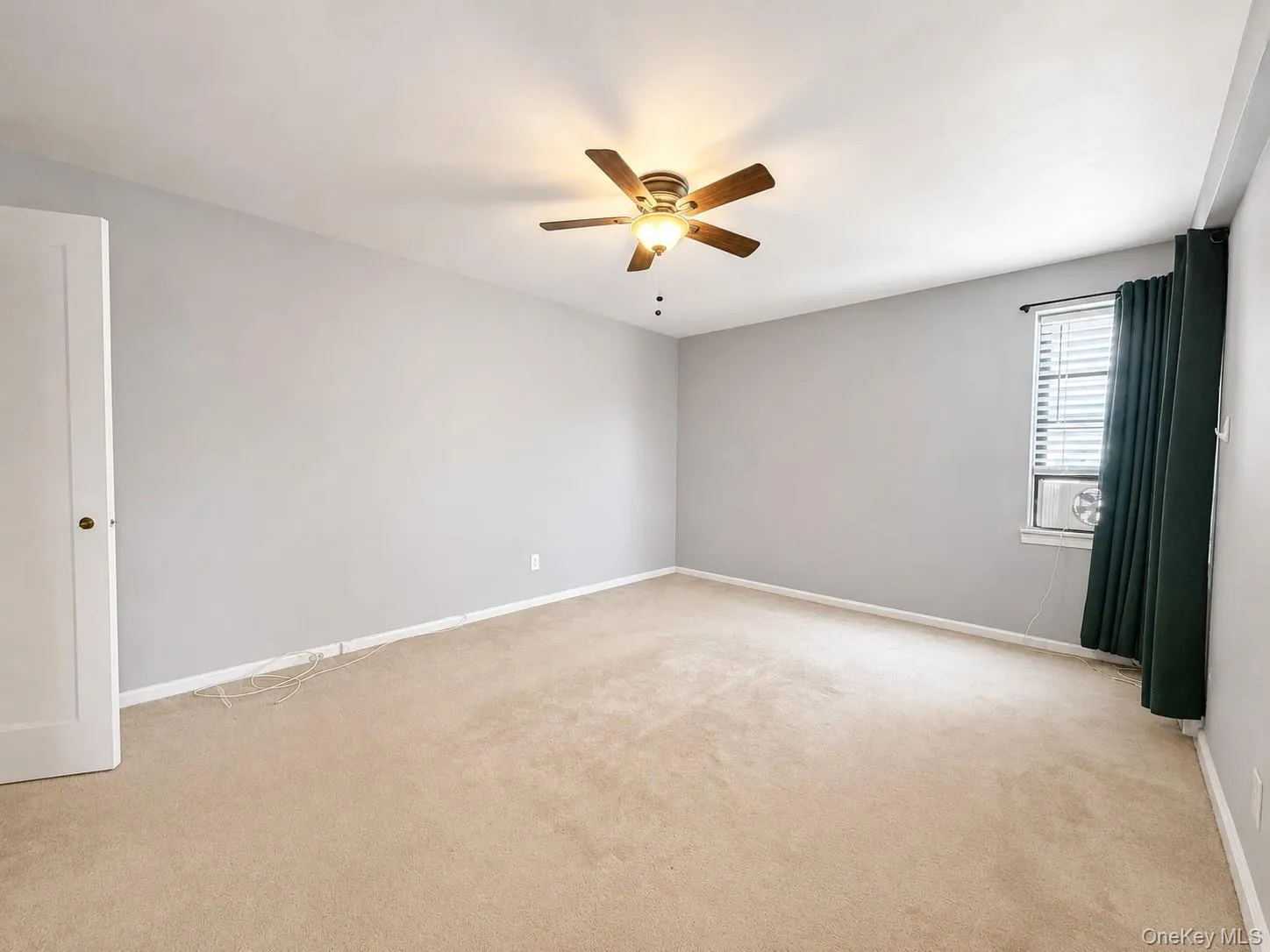Expansive room featuring neutral-toned carpet, light gray wall finishes, a ceiling fan with integrated lighting, a window with horizontal blinds, and a white interior door with a brass-toned knob Expansive room featuring neutral-toned carpet, light gray wall finishes, a ceiling fan with integrated lighting, a window with horizontal blinds, and a white interior door with a brass-toned knob