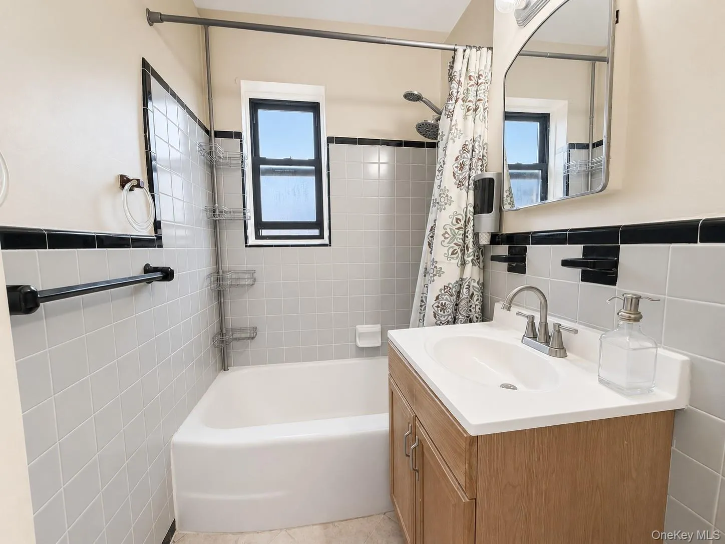 Bathroom featuring light-toned square tile walls with a black accent border, a white bathtub, and a window with dark framing Bathroom featuring light-toned square tile walls with a black accent border, a white bathtub, and a window with dark framing