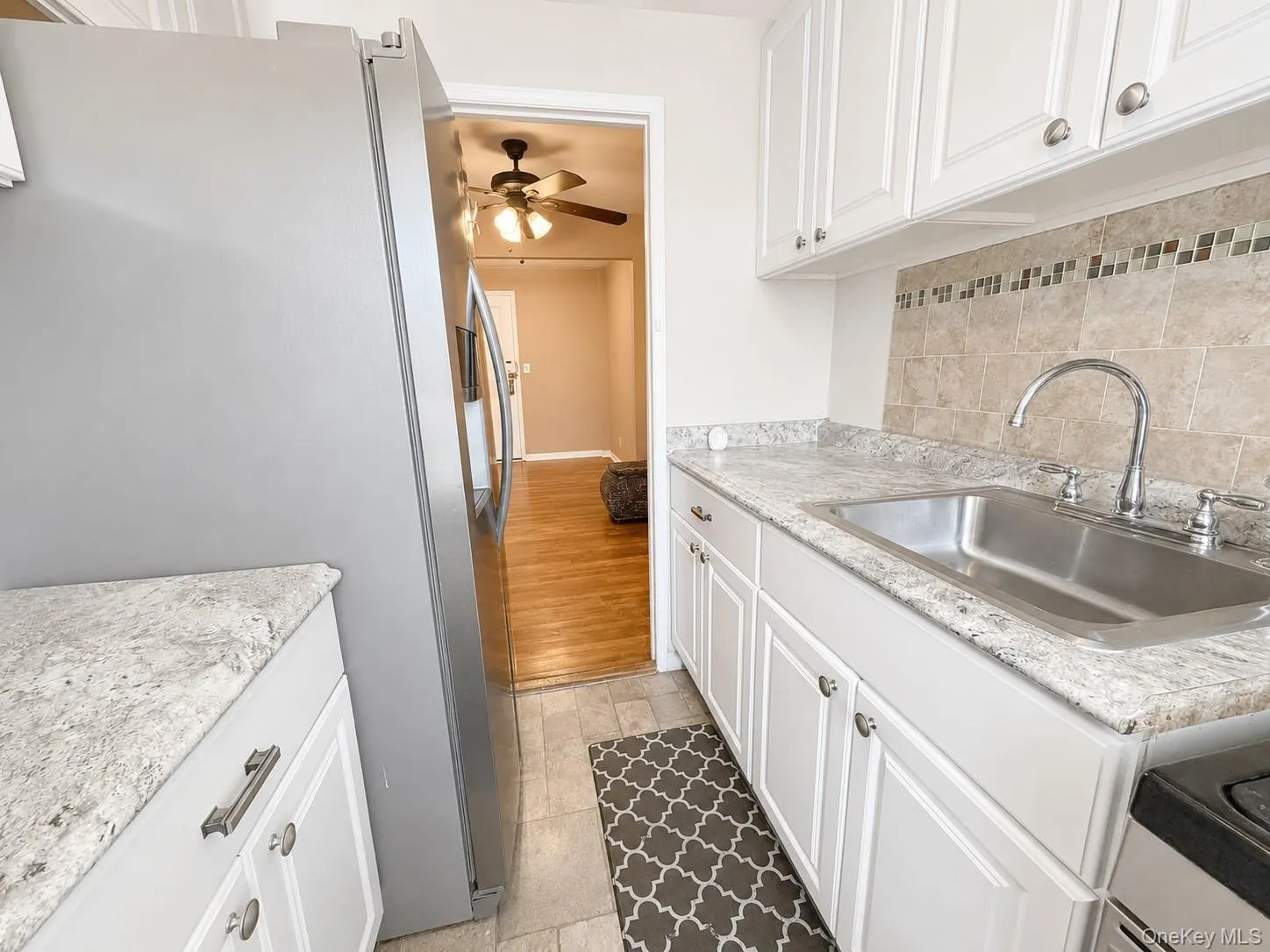 Kitchen featuring white shaker-style cabinetry with brushed nickel hardware, granite-look countertops, a stainless steel undermount sink with a chrome faucet, and a stone tile backsplash with decorative mosaic accents Kitchen featuring white shaker-style cabinetry with brushed nickel hardware, granite-look countertops, a stainless steel undermount sink with a chrome faucet, and a stone tile backsplash with decorative mosaic accents