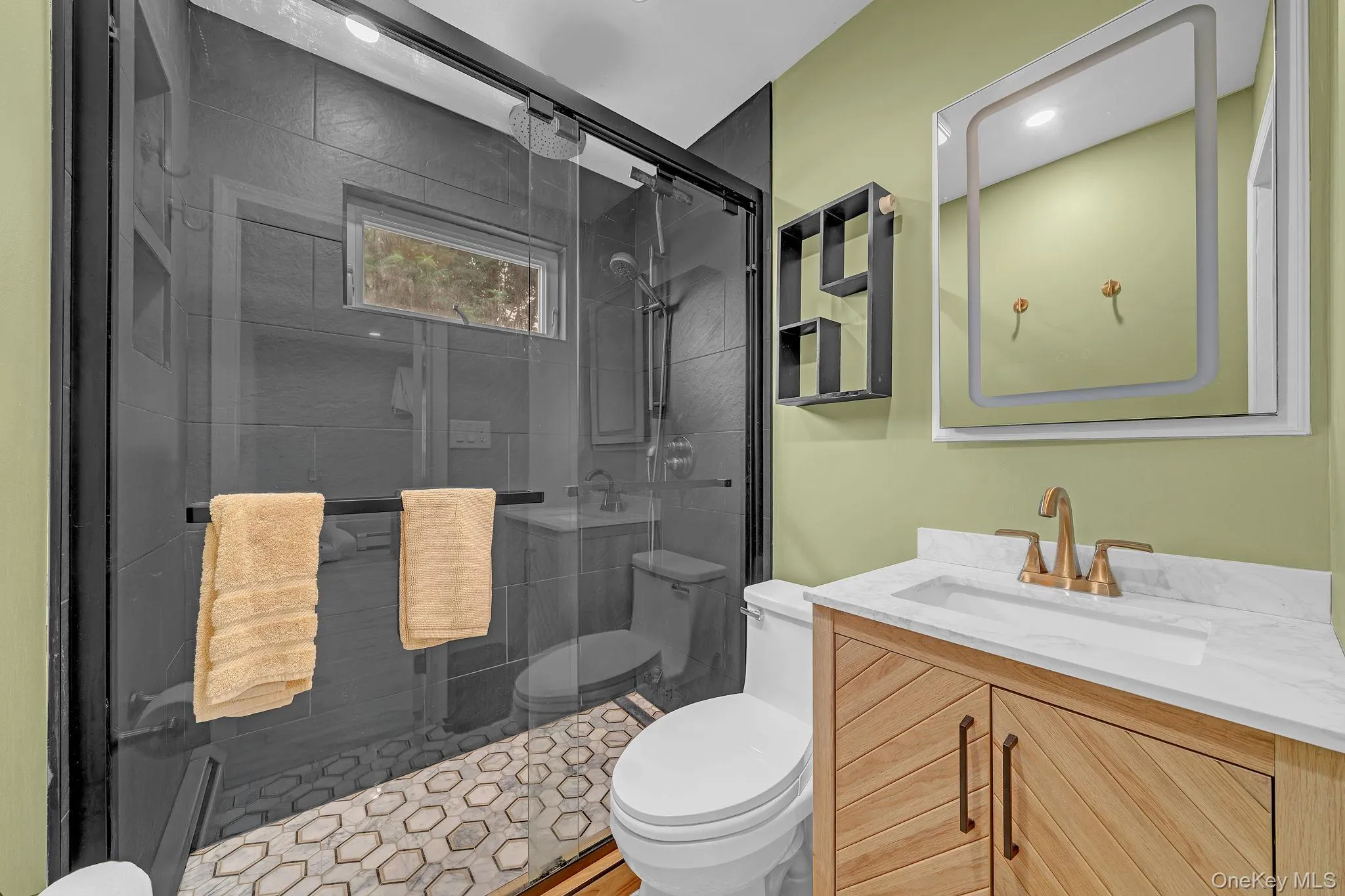 Bathroom featuring a glass-enclosed shower with black tile surround, a wood-finish vanity with a white countertop and gold-tone faucet, a lighted mirror, and a wall-mounted shelving unit Bathroom featuring a glass-enclosed shower with black tile surround, a wood-finish vanity with a white countertop and gold-tone faucet, a lighted mirror, and a wall-mounted shelving unit