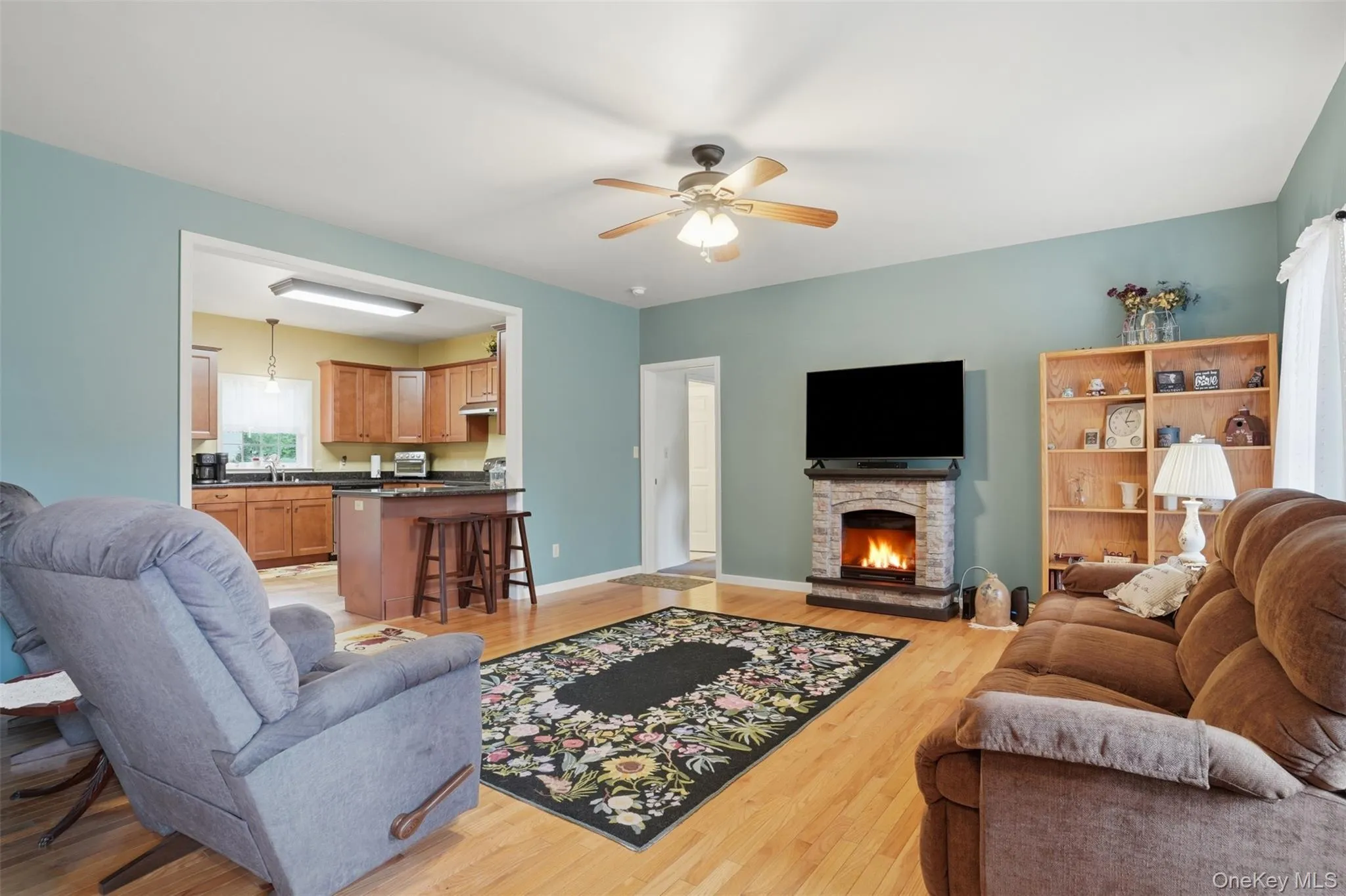 Open-concept living area featuring wood-finish flooring, a stone-look fireplace hearth, and a ceiling fan Open-concept living area featuring wood-finish flooring, a stone-look fireplace hearth, and a ceiling fan