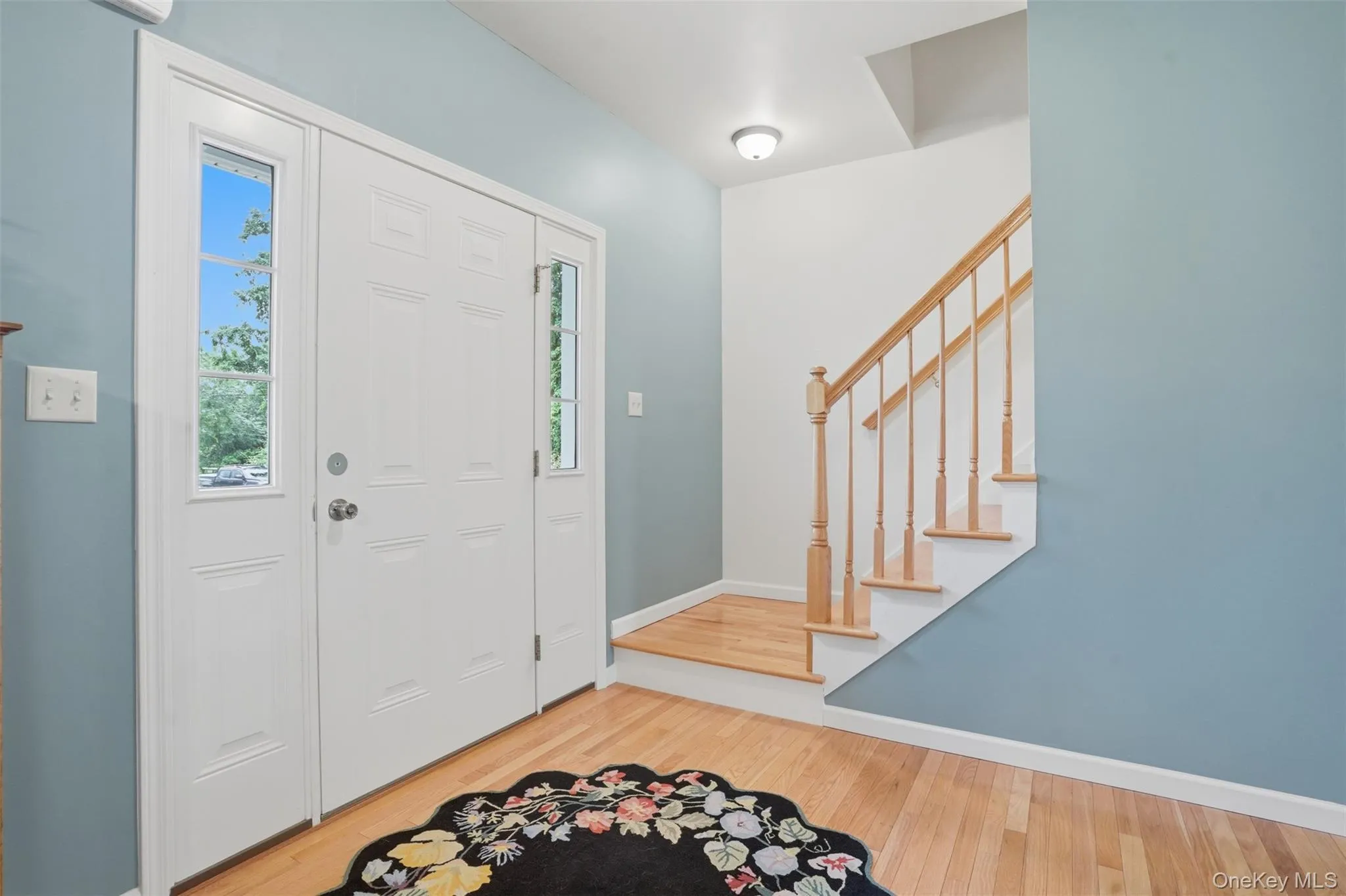 Entry foyer featuring a paneled door with sidelights, wood-finish flooring, a wood staircase, and a flush-mount ceiling light fixture Entry foyer featuring a paneled door with sidelights, wood-finish flooring, a wood staircase, and a flush-mount ceiling light fixture