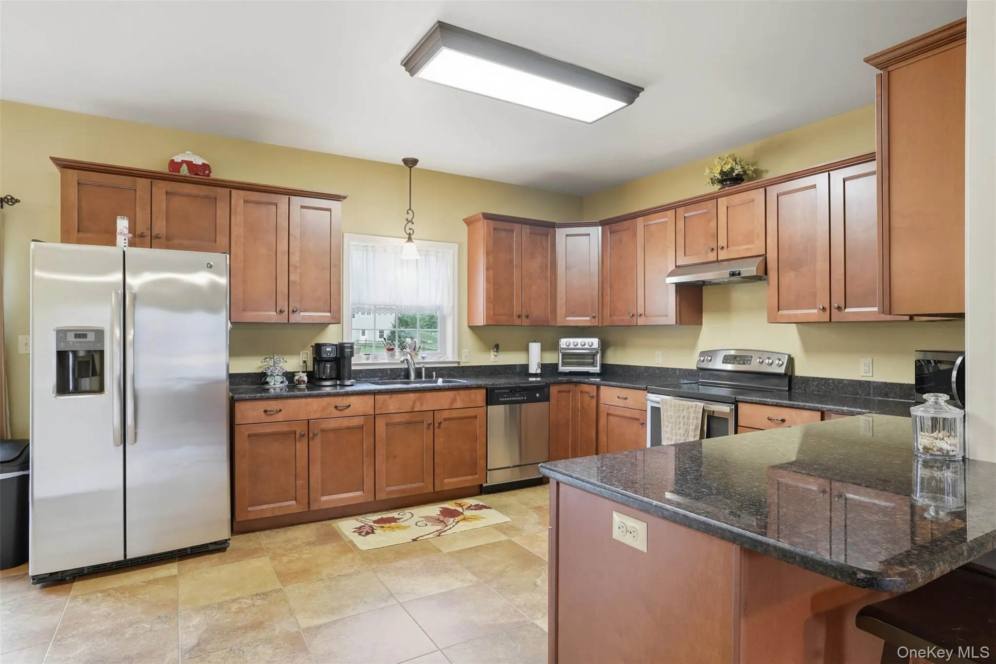 Kitchen featuring tile flooring, wood cabinetry, dark countertops, a stainless steel refrigerator, and a built-in kitchen island Kitchen featuring tile flooring, wood cabinetry, dark countertops, a stainless steel refrigerator, and a built-in kitchen island