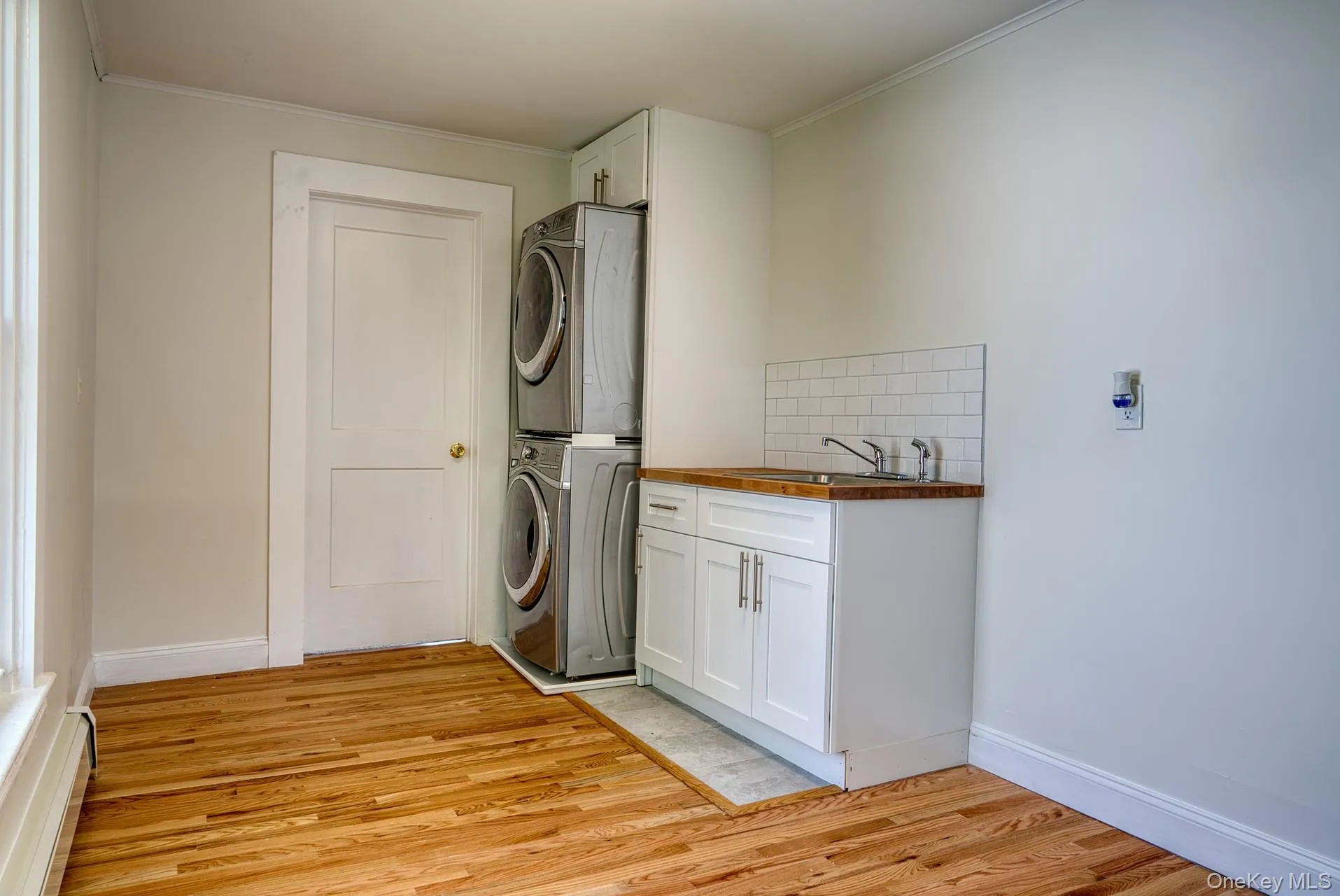 Washroom with stacked washing machine and dryer, cabinet space, light wood-type flooring, ornamental molding, and baseboards Washroom with stacked washing machine and dryer, cabinet space, light wood-type flooring, ornamental molding, and baseboards