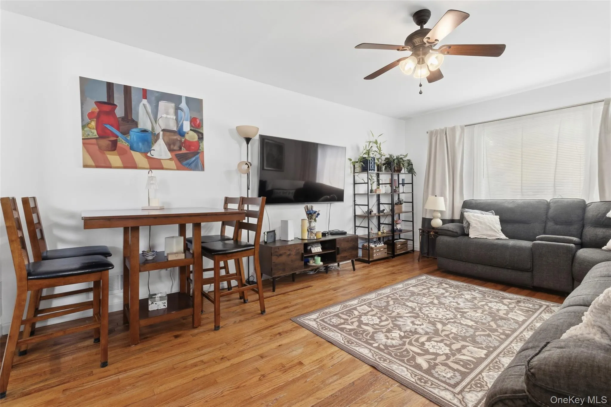 Living area featuring wood-finish flooring, a ceiling fan with integrated lighting, and a large window with privacy blinds Living area featuring wood-finish flooring, a ceiling fan with integrated lighting, and a large window with privacy blinds