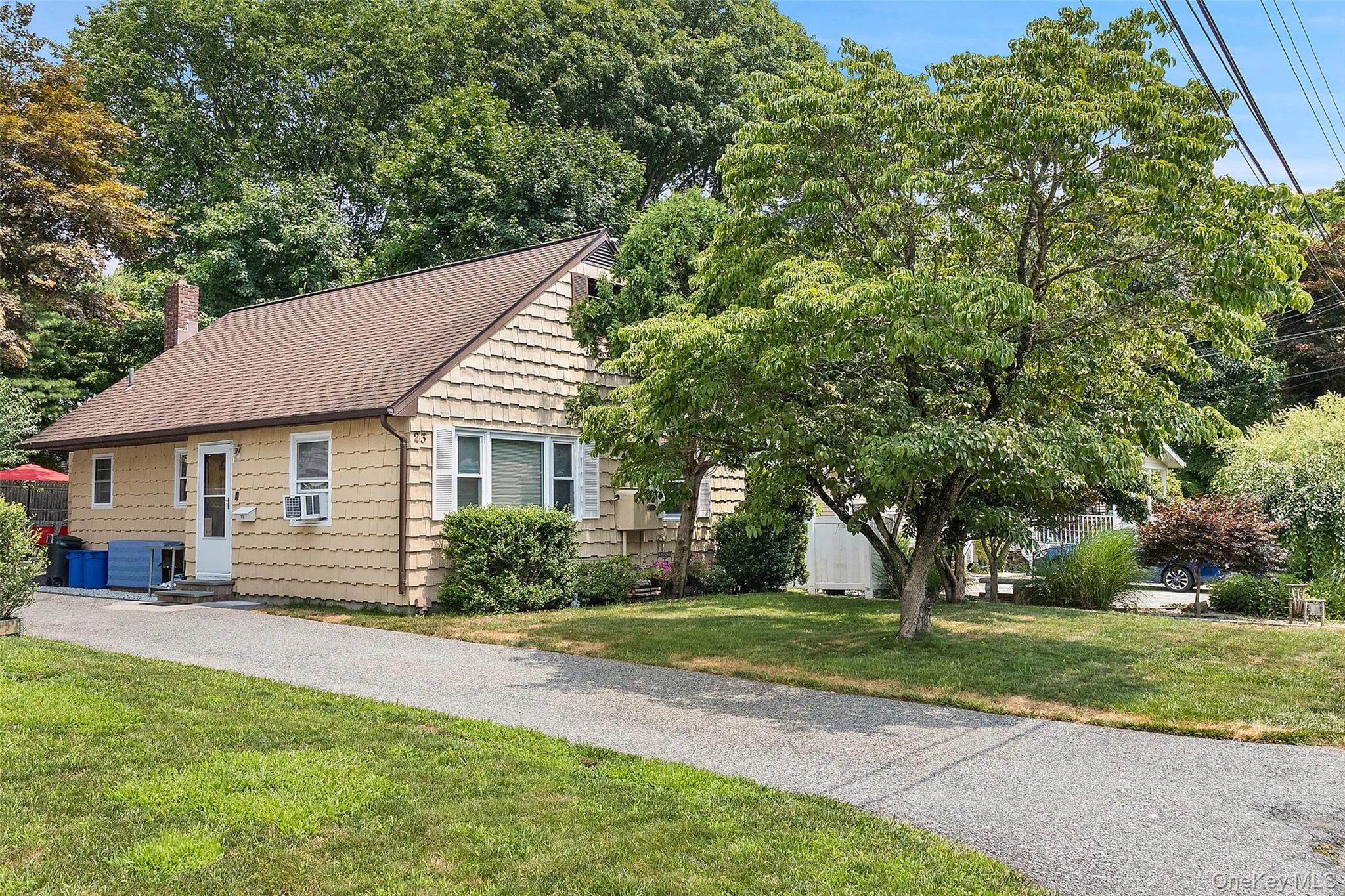 Beige shake siding exterior with a brown shingle roof, a paved driveway, and mature landscaping Beige shake siding exterior with a brown shingle roof, a paved driveway, and mature landscaping