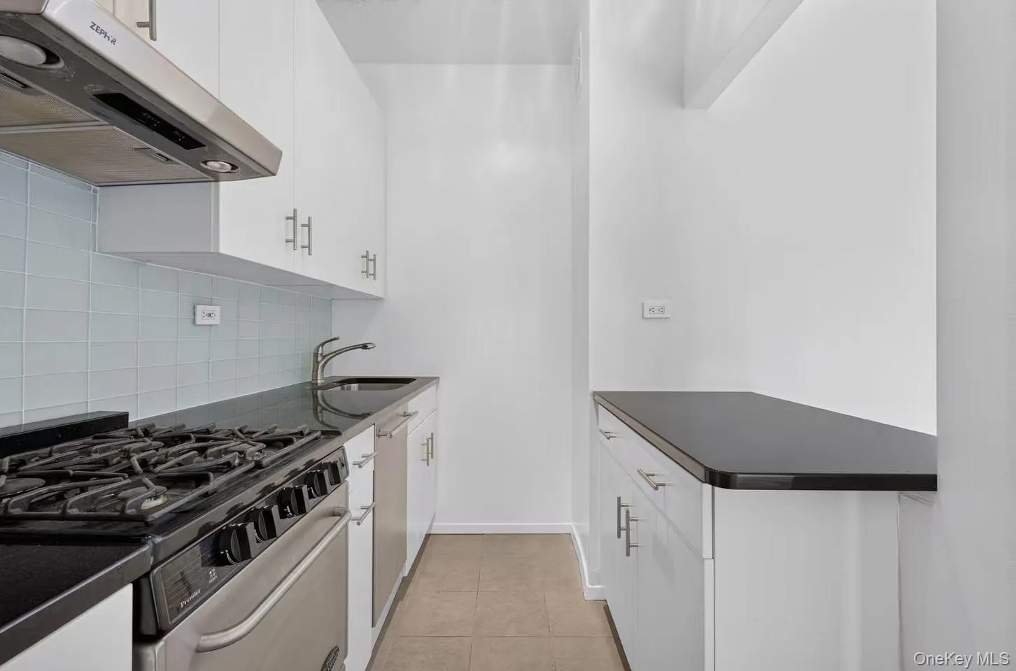 Galley kitchen featuring white cabinetry, dark countertops, a gas range with stainless steel oven, an undermount sink, and a light blue tiled backsplash Galley kitchen featuring white cabinetry, dark countertops, a gas range with stainless steel oven, an undermount sink, and a light blue tiled backsplash