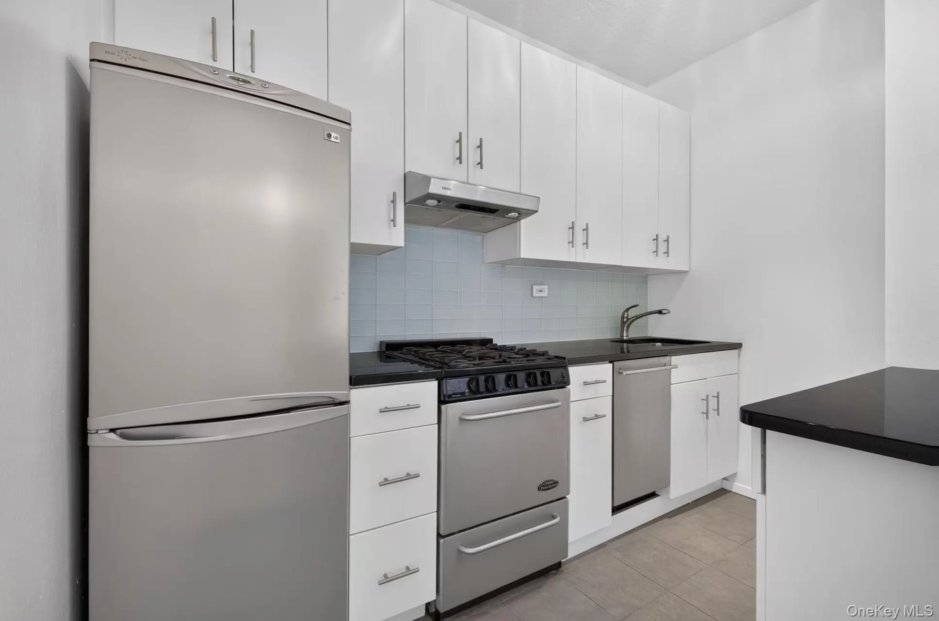 Streamlined kitchen featuring white cabinetry, dark countertops, stainless steel appliances, a tile backsplash, and wood-finish flooring Streamlined kitchen featuring white cabinetry, dark countertops, stainless steel appliances, a tile backsplash, and wood-finish flooring