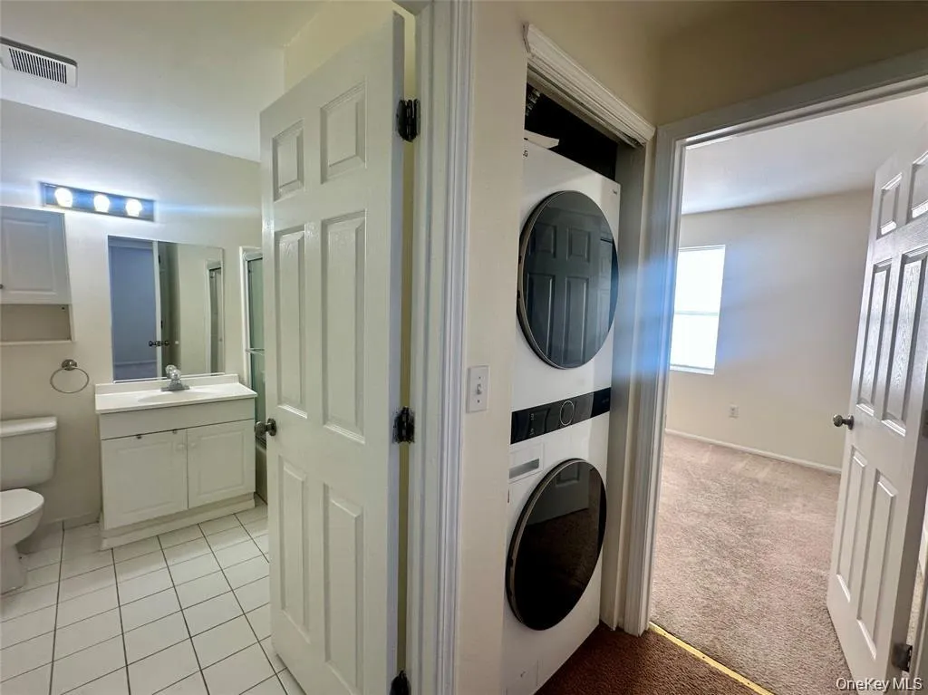 Stacked laundry appliances, white panel doors, vanity with integrated sink, wall-mounted mirror, and tile flooring Stacked laundry appliances, white panel doors, vanity with integrated sink, wall-mounted mirror, and tile flooring