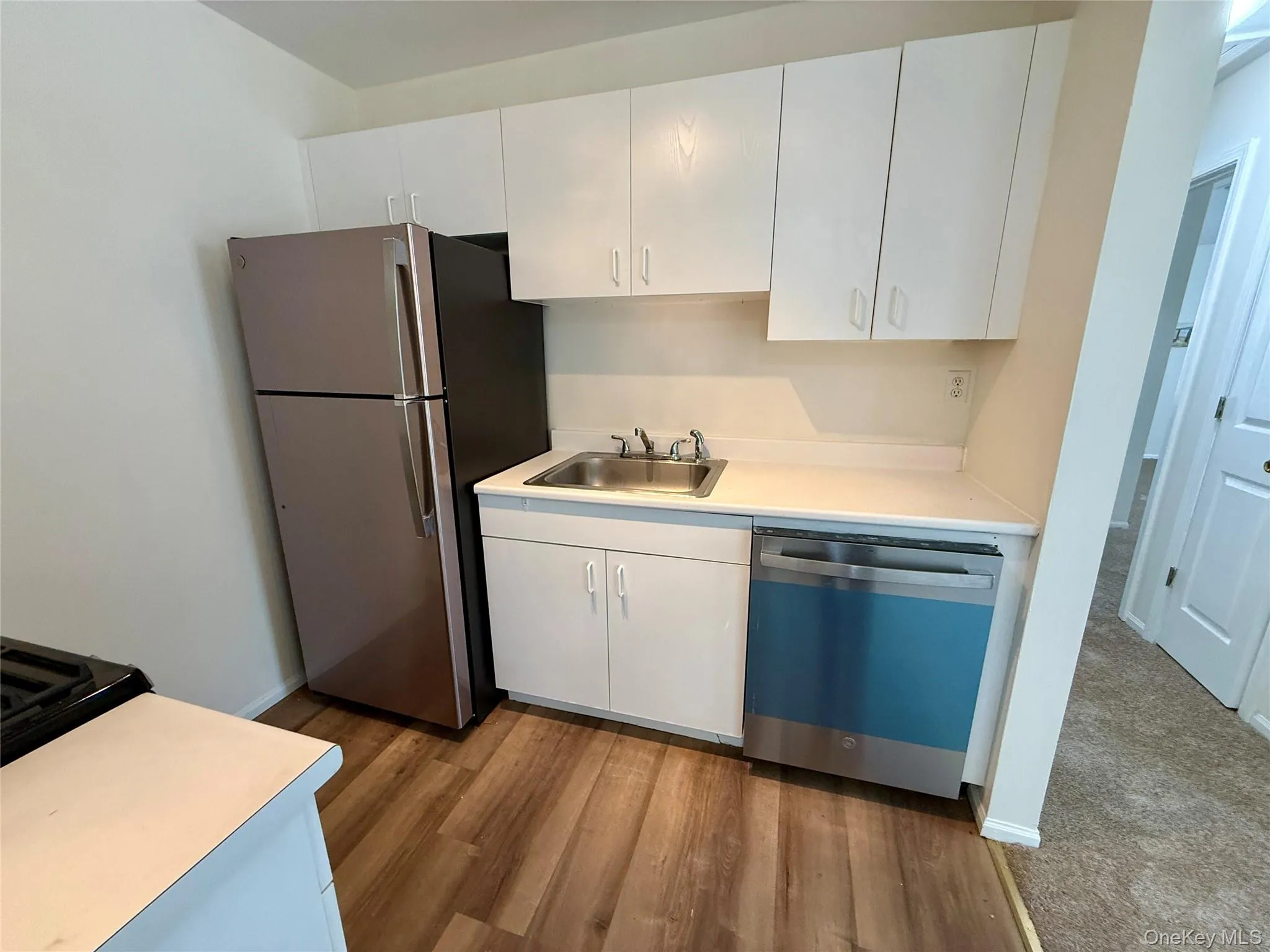 White cabinetry with brushed nickel hardware, stainless steel sink, wood-finish flooring, and white countertops White cabinetry with brushed nickel hardware, stainless steel sink, wood-finish flooring, and white countertops