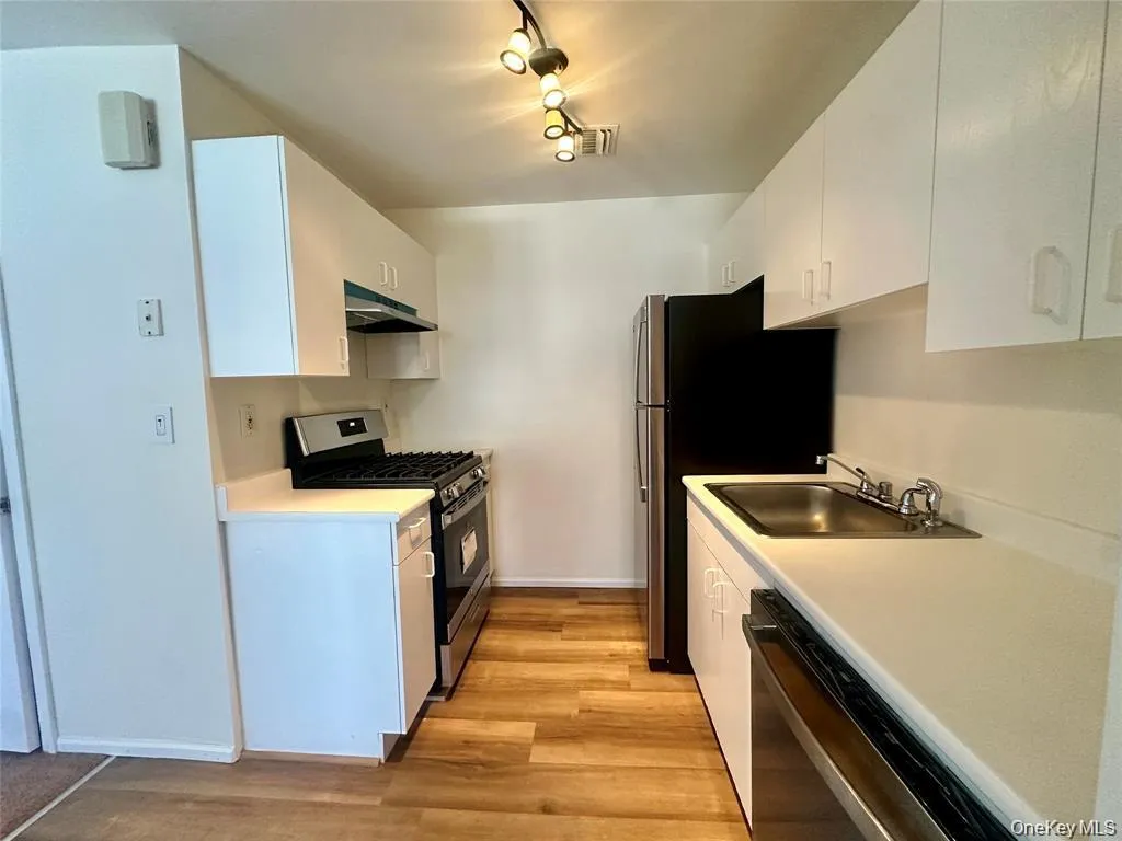 Galley kitchen with wood-finish flooring, white cabinetry, stainless steel appliances, and track lighting Galley kitchen with wood-finish flooring, white cabinetry, stainless steel appliances, and track lighting