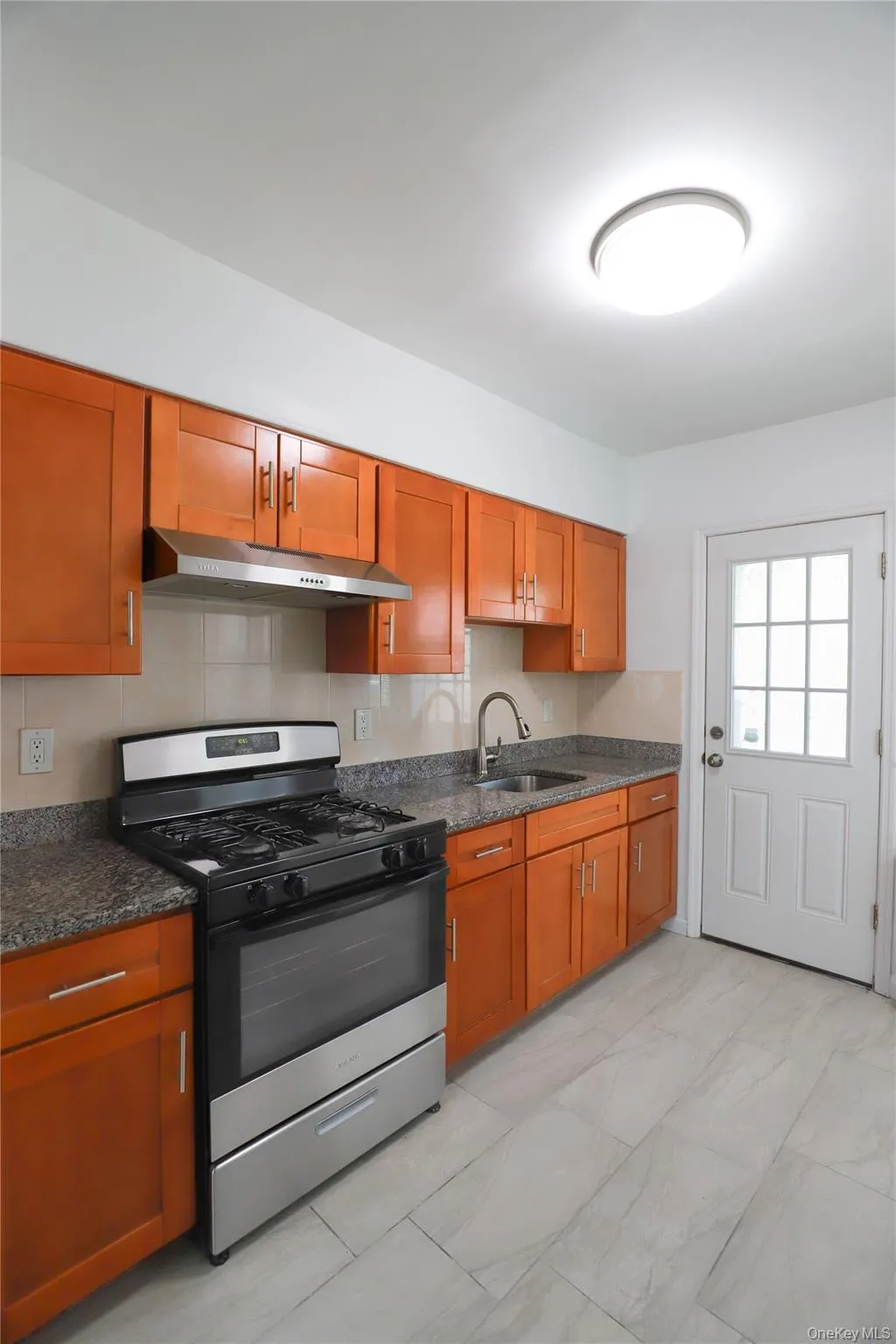 Kitchen featuring wood-finish cabinetry, speckled countertops, stainless steel range with matching hood, and light-toned tile flooring Kitchen featuring wood-finish cabinetry, speckled countertops, stainless steel range with matching hood, and light-toned tile flooring