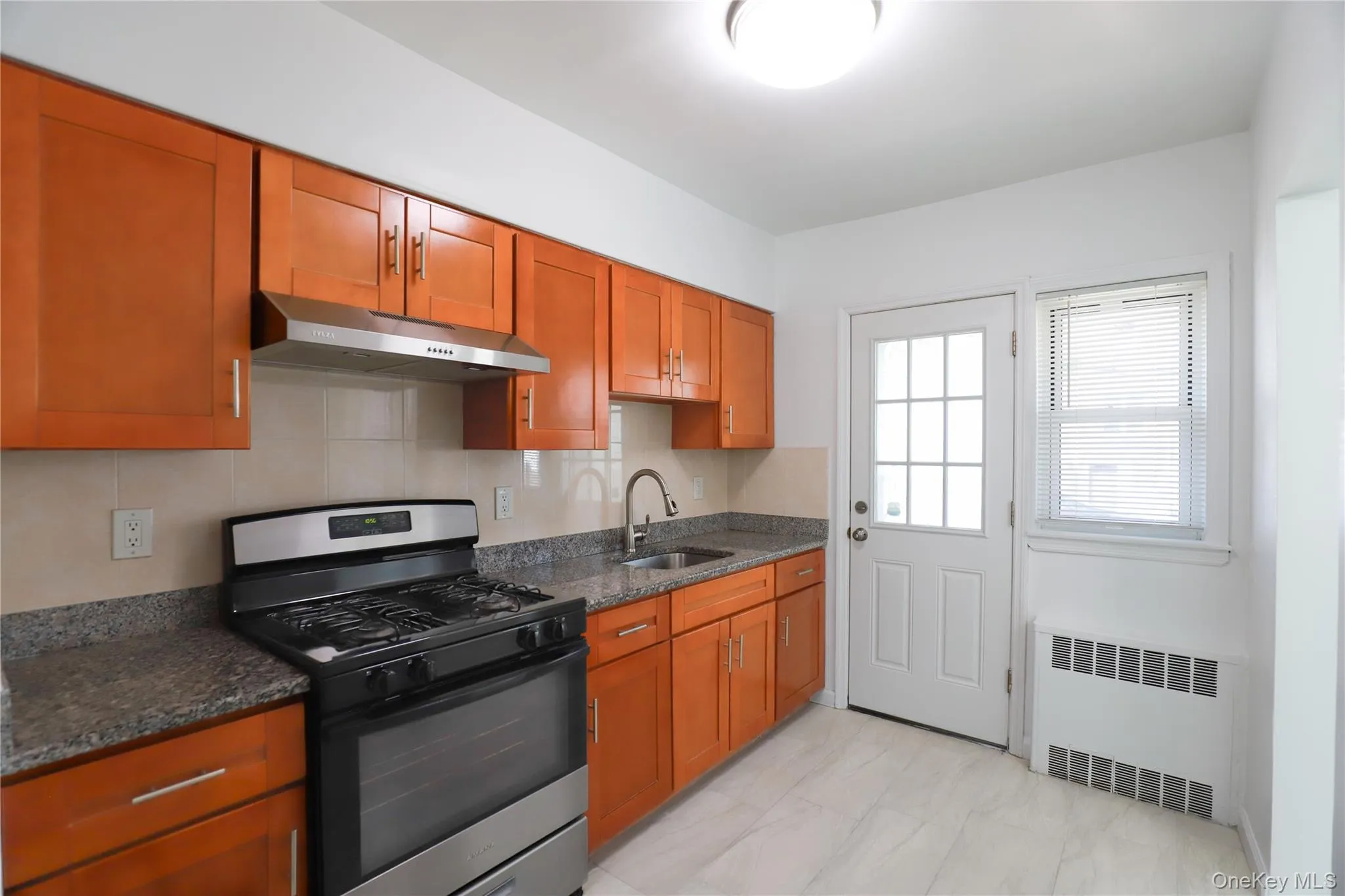 Kitchen featuring wood-finish cabinetry, speckled countertops, stainless steel range with matching hood, and light-toned tile flooring Kitchen featuring wood-finish cabinetry, speckled countertops, stainless steel range with matching hood, and light-toned tile flooring