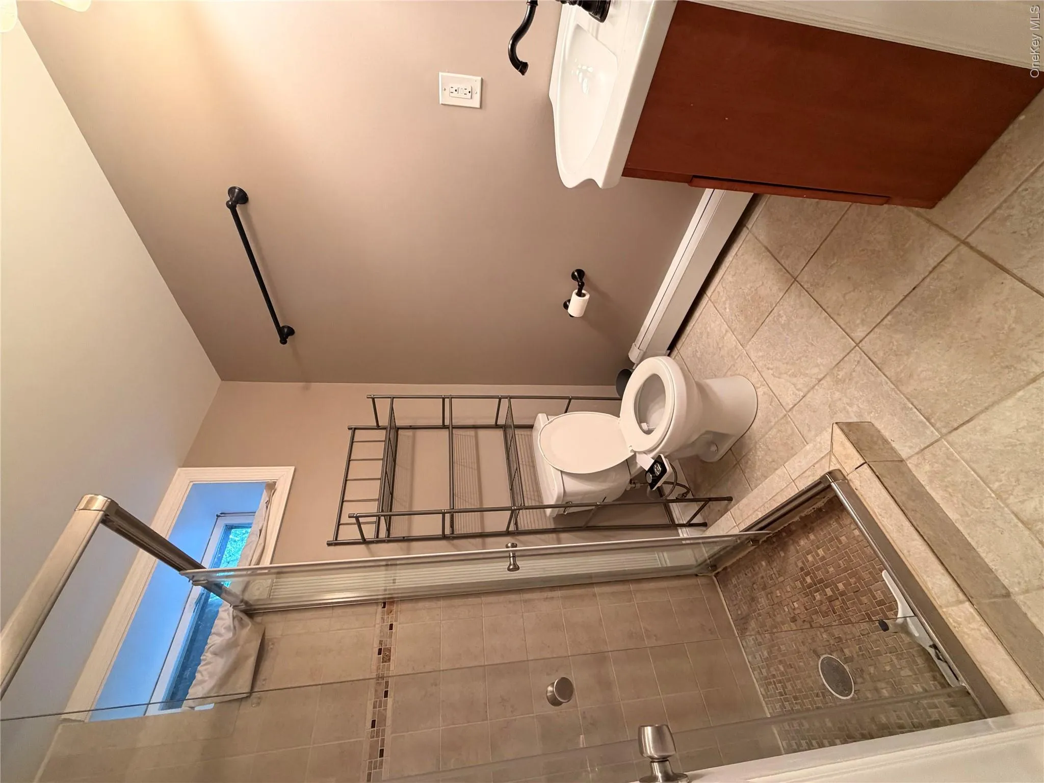 Bathroom featuring a glass-enclosed shower with tile surround and mosaic floor, a single vanity with a white basin and dark-toned cabinet, and neutral tile flooring Bathroom featuring a glass-enclosed shower with tile surround and mosaic floor, a single vanity with a white basin and dark-toned cabinet, and neutral tile flooring