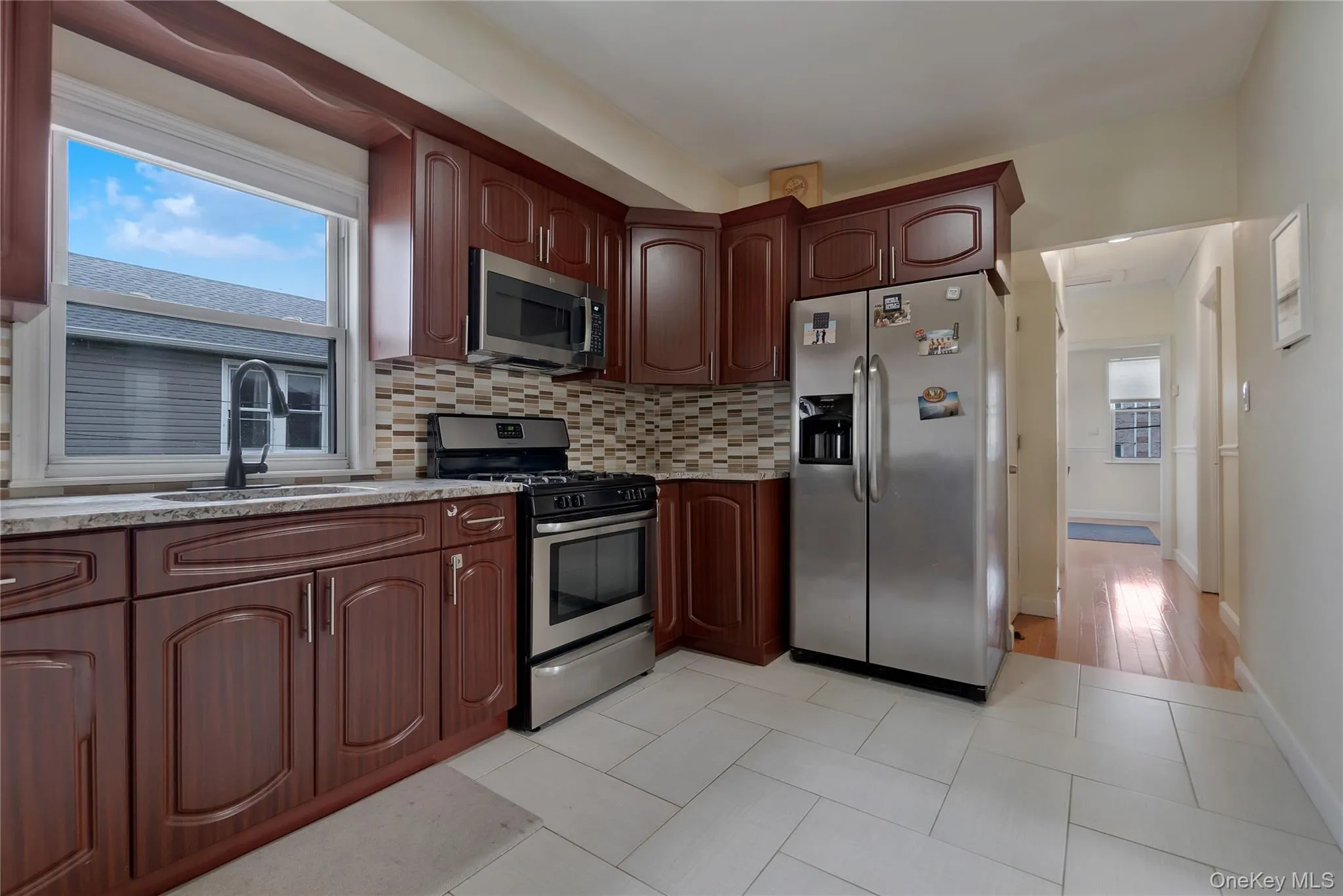 Kitchen featuring wood-finish cabinetry, stainless steel appliances, a multi-tone tile backsplash, and light-toned tile flooring Kitchen featuring wood-finish cabinetry, stainless steel appliances, a multi-tone tile backsplash, and light-toned tile flooring