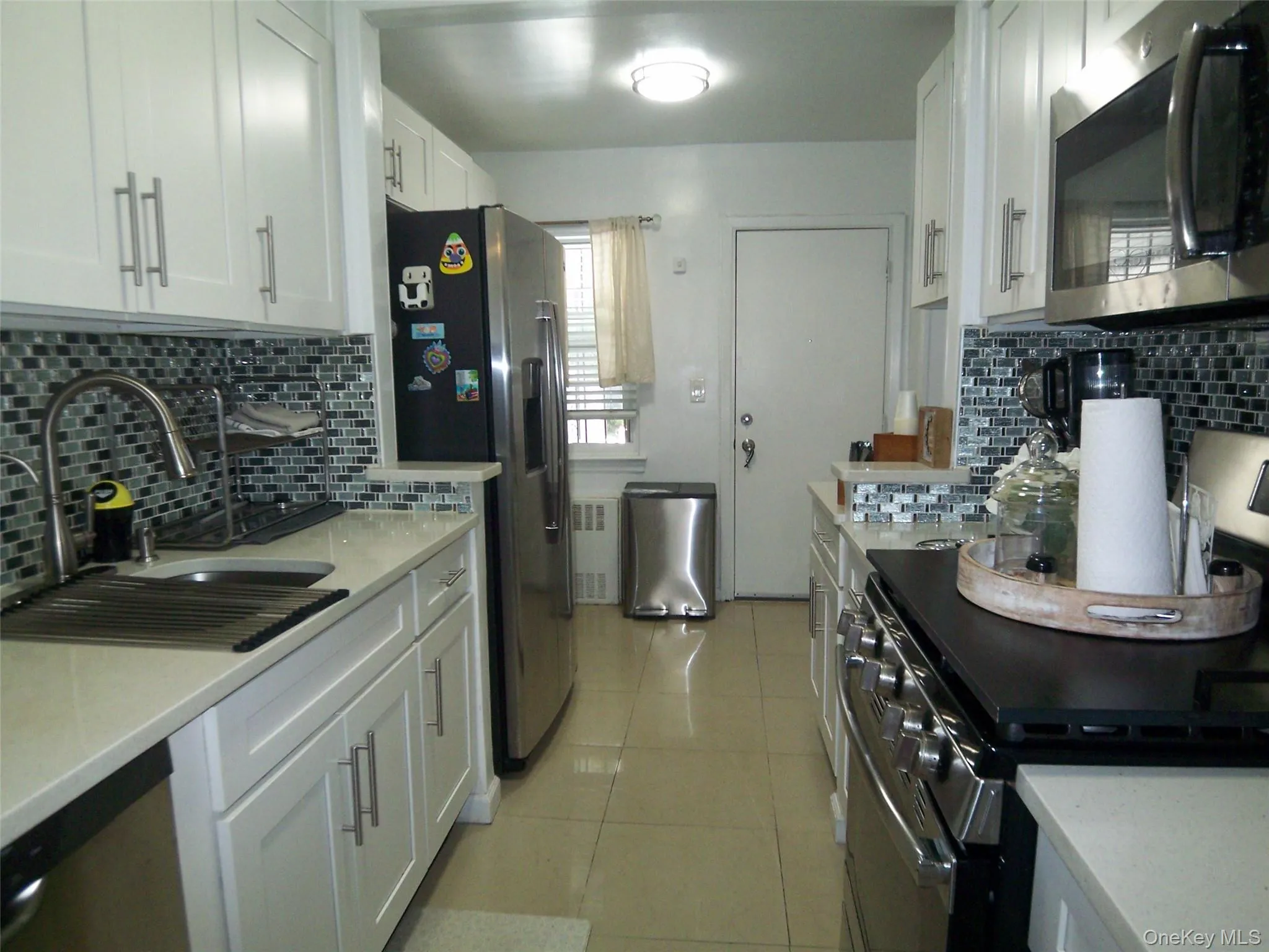 Galley kitchen featuring white cabinetry with brushed nickel pulls, light-toned countertops, and a mosaic tile backsplash Galley kitchen featuring white cabinetry with brushed nickel pulls, light-toned countertops, and a mosaic tile backsplash