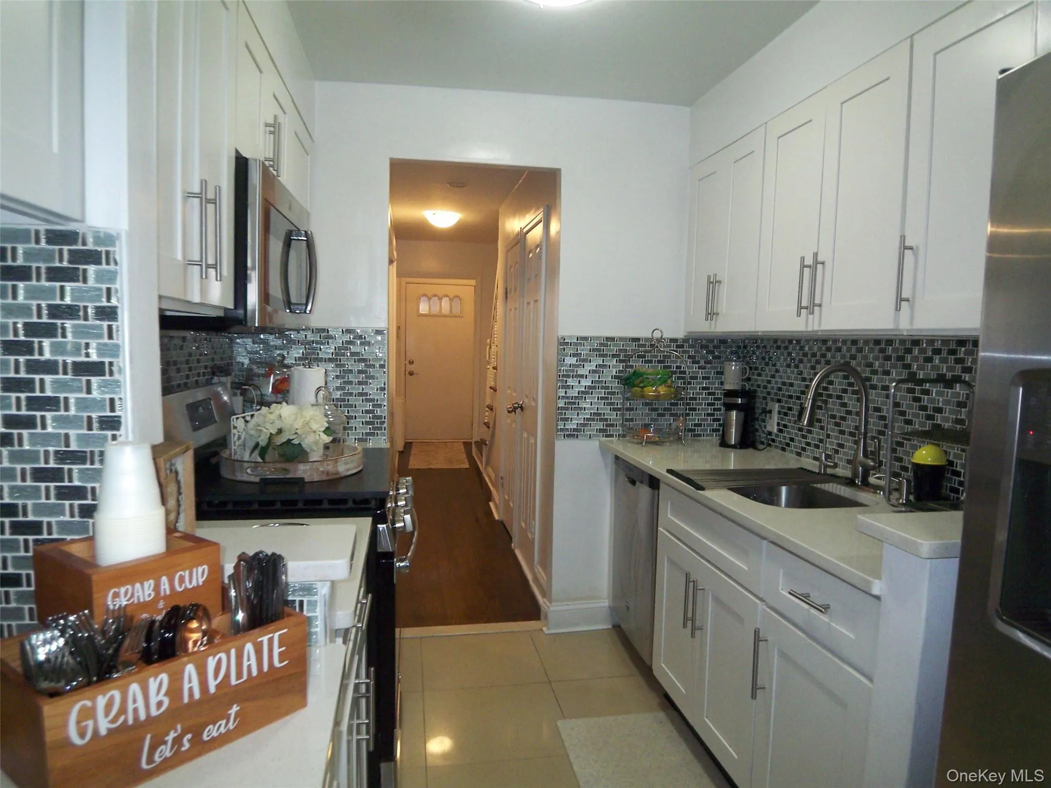 Galley kitchen featuring white shaker cabinetry, stainless steel appliances, a tiled backsplash, and light-tone countertops Galley kitchen featuring white shaker cabinetry, stainless steel appliances, a tiled backsplash, and light-tone countertops