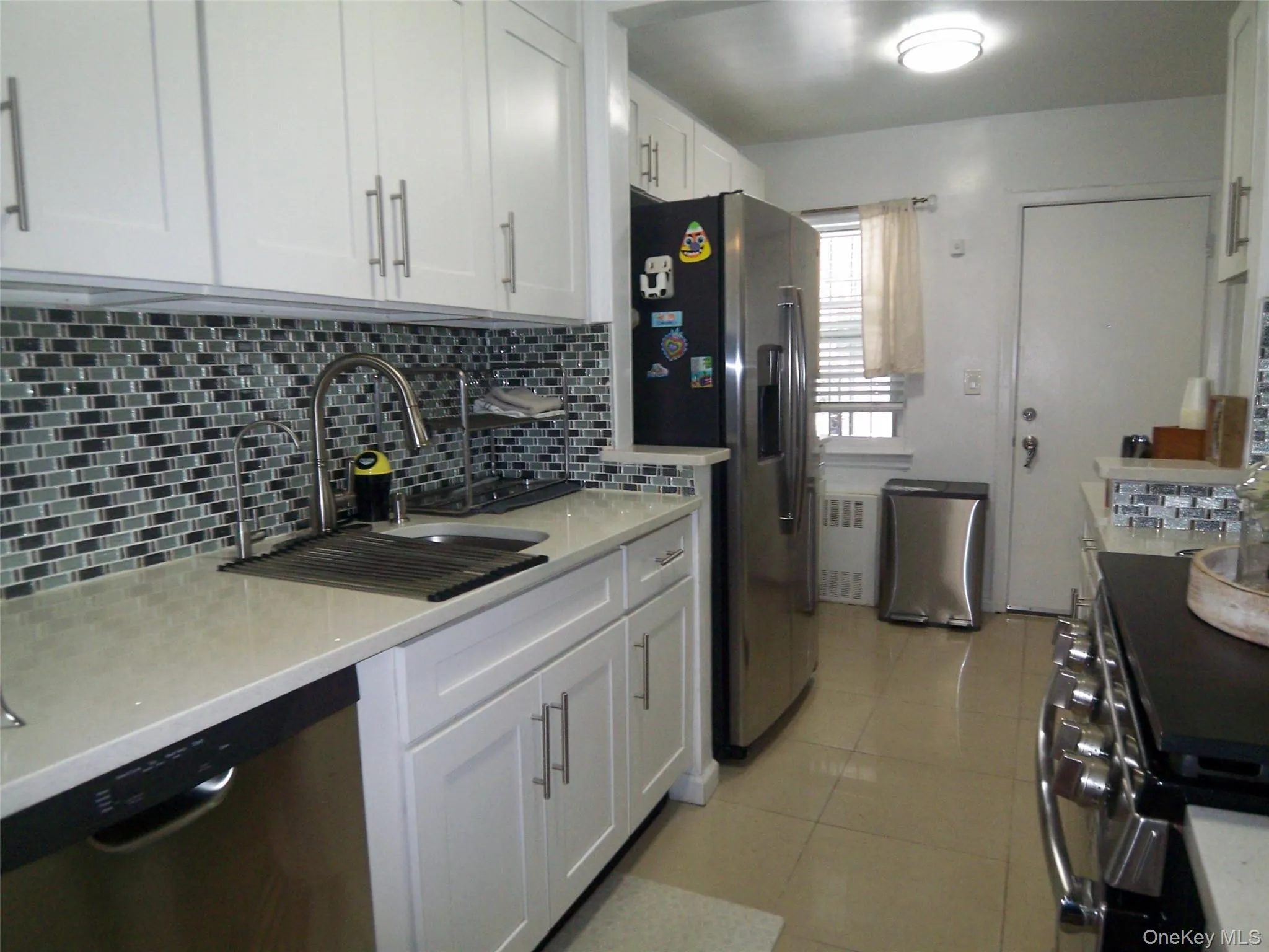 Modern kitchen featuring white shaker cabinetry, light-toned countertops, a mosaic tile backsplash, stainless steel appliances, and tile flooring Modern kitchen featuring white shaker cabinetry, light-toned countertops, a mosaic tile backsplash, stainless steel appliances, and tile flooring