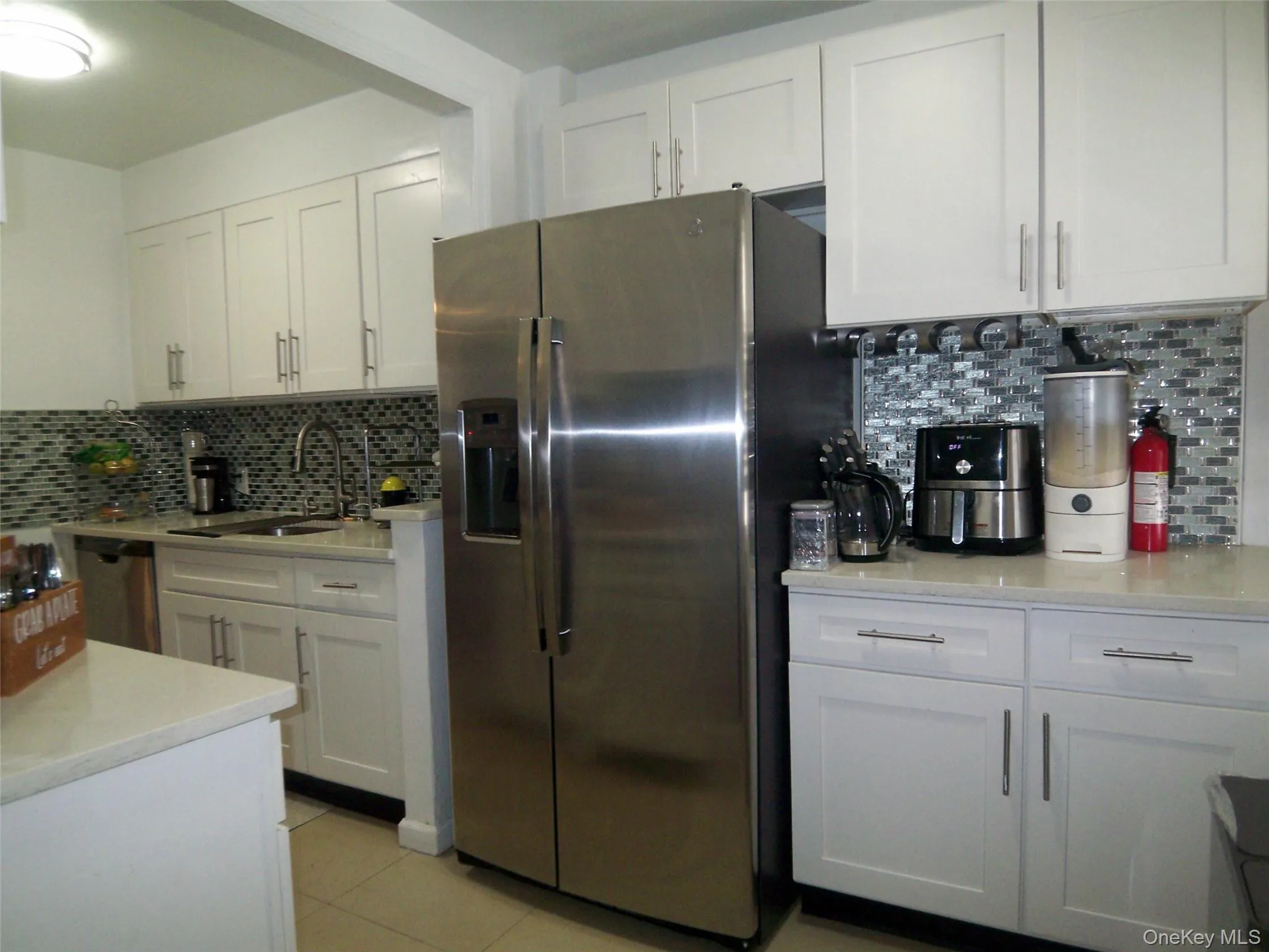Modern kitchen featuring a stainless steel side-by-side refrigerator, white shaker-style cabinetry, light-toned countertops, a mosaic tile backsplash, and a stainless steel undermount sink Modern kitchen featuring a stainless steel side-by-side refrigerator, white shaker-style cabinetry, light-toned countertops, a mosaic tile backsplash, and a stainless steel undermount sink
