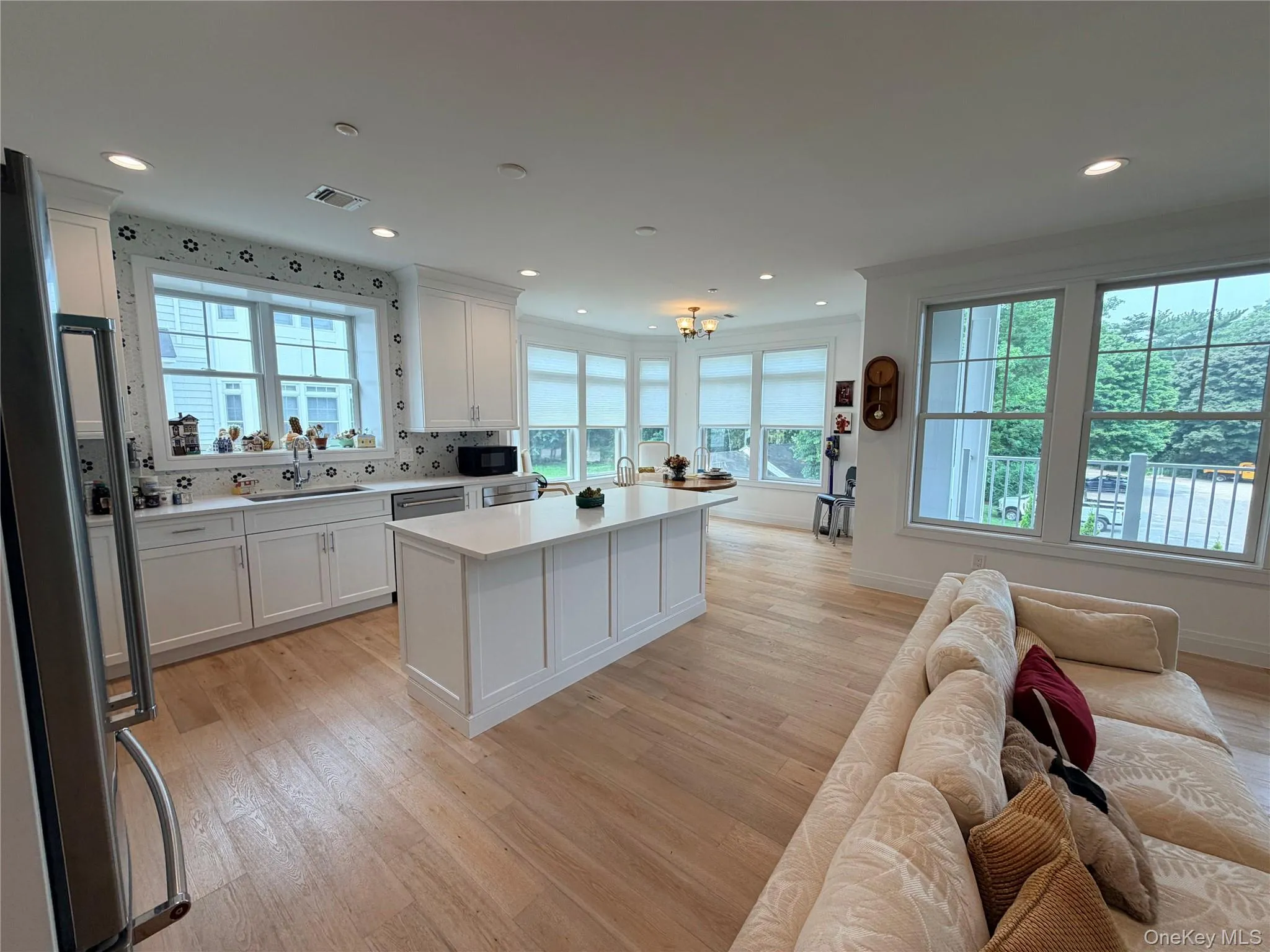 Spacious open-concept kitchen and living area featuring wood-finish flooring, white cabinetry, a large kitchen island with a light-toned countertop, recessed lighting, and multiple windows with roller shades Spacious open-concept kitchen and living area featuring wood-finish flooring, white cabinetry, a large kitchen island with a light-toned countertop, recessed lighting, and multiple windows with roller shades