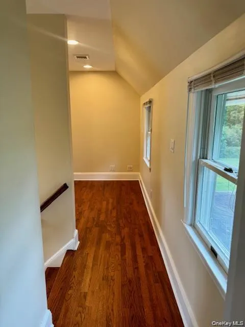 Hallway featuring wood-finish flooring, two windows with blinds, recessed lighting, and a partial staircase with a wood handrail Hallway featuring wood-finish flooring, two windows with blinds, recessed lighting, and a partial staircase with a wood handrail