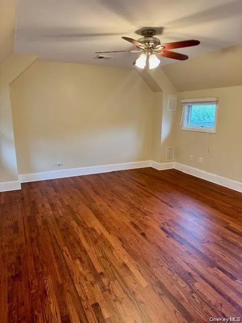 Hardwood flooring spans this expansive room featuring a ceiling fan with integrated lighting, a window with exterior visibility, and sloped ceiling details Hardwood flooring spans this expansive room featuring a ceiling fan with integrated lighting, a window with exterior visibility, and sloped ceiling details