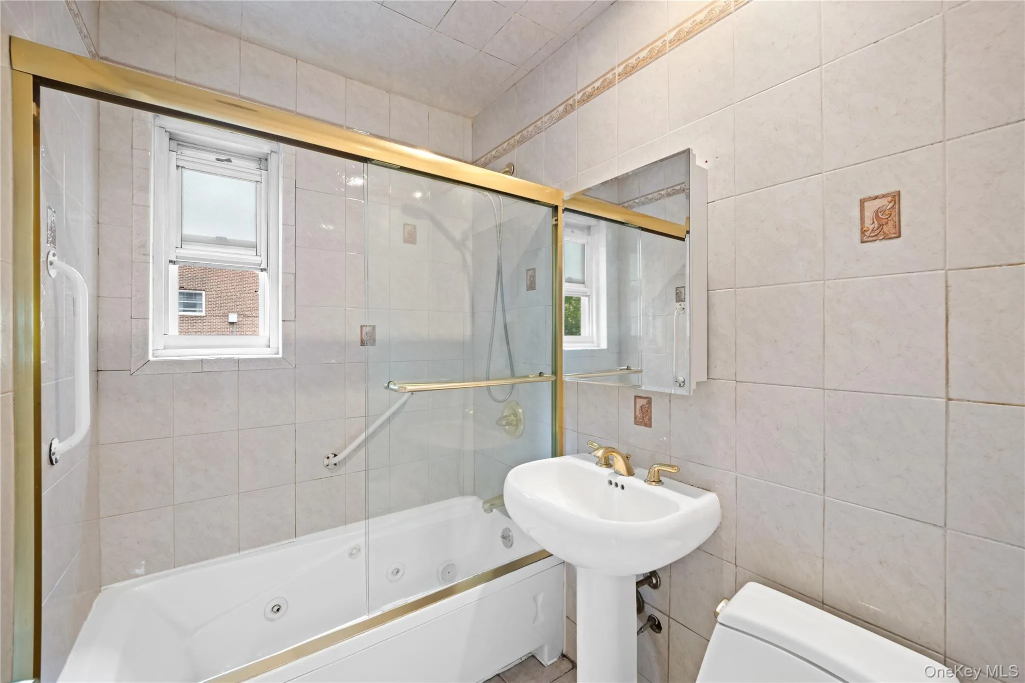 Full bathroom featuring a pedestal sink with gold-tone fixtures, a jetted bathtub with a sliding glass enclosure, and tiled walls Full bathroom featuring a pedestal sink with gold-tone fixtures, a jetted bathtub with a sliding glass enclosure, and tiled walls