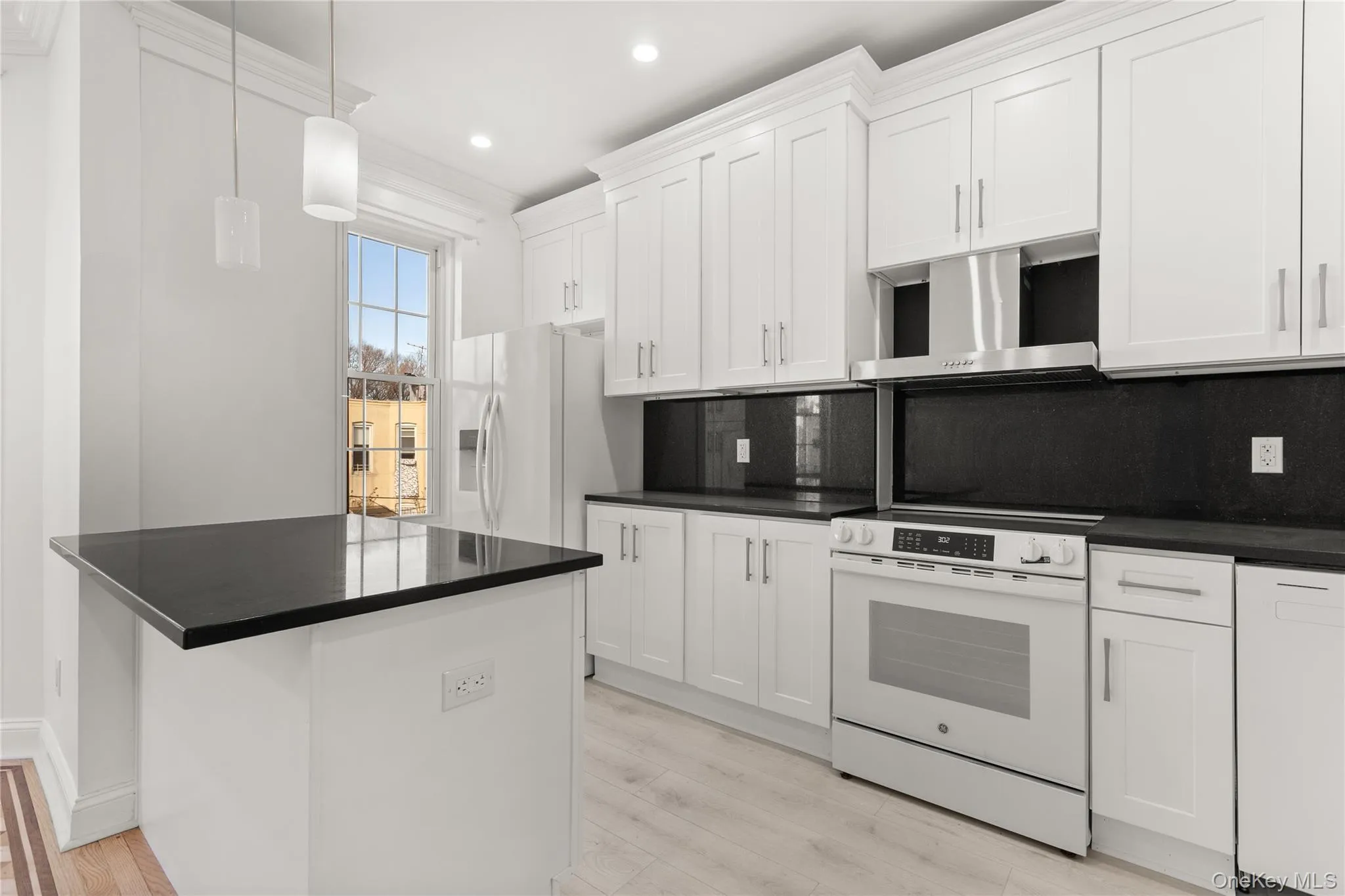 Contemporary kitchen featuring white shaker cabinetry, dark countertops, stainless steel range hood, built-in oven, and wood-finish flooring Contemporary kitchen featuring white shaker cabinetry, dark countertops, stainless steel range hood, built-in oven, and wood-finish flooring