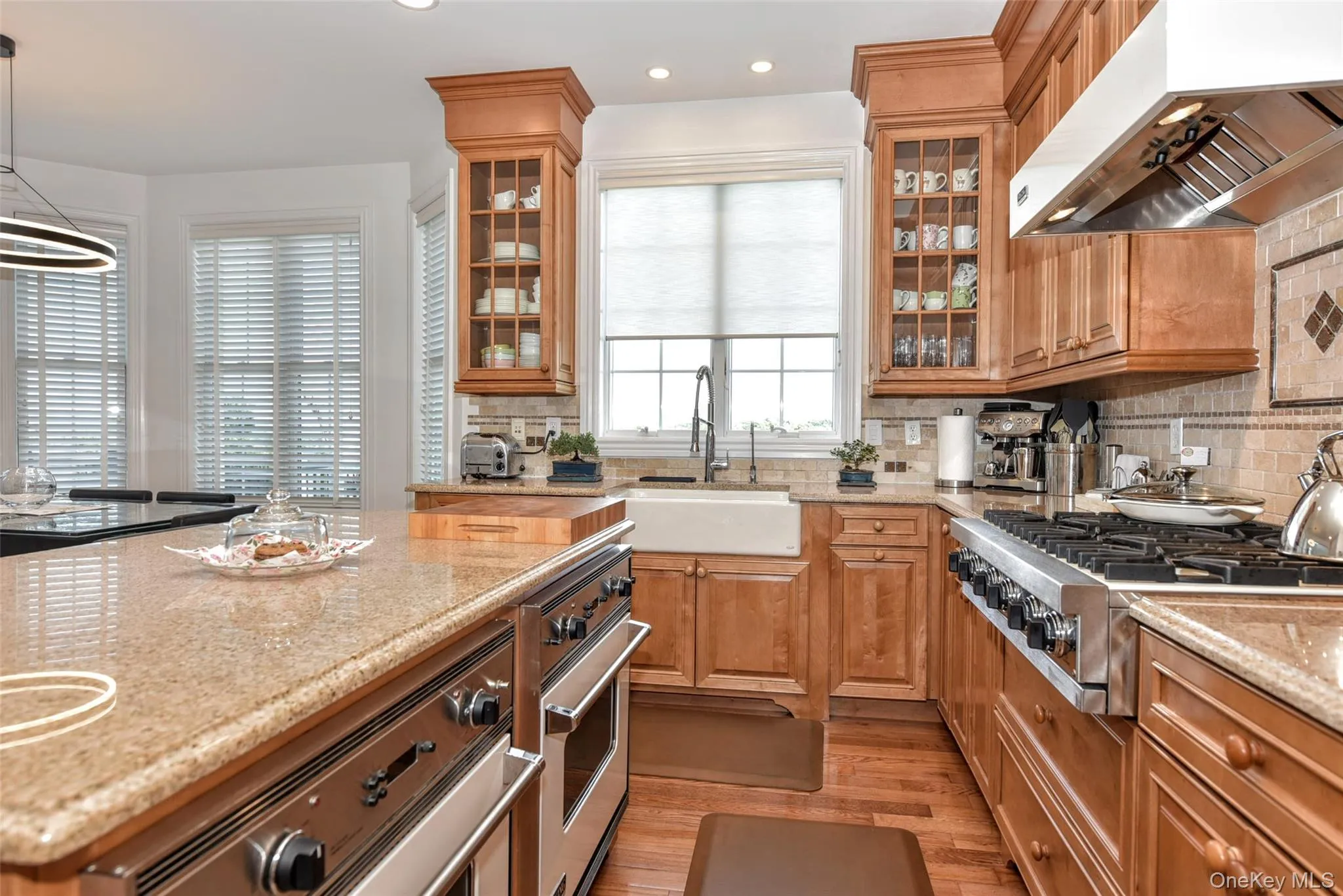 Gourmet kitchen featuring a large central island with integrated oven, a built-in range with an overhead exhaust hood, wood-finish cabinetry, a farmhouse sink, and polished countertops Gourmet kitchen featuring a large central island with integrated oven, a built-in range with an overhead exhaust hood, wood-finish cabinetry, a farmhouse sink, and polished countertops