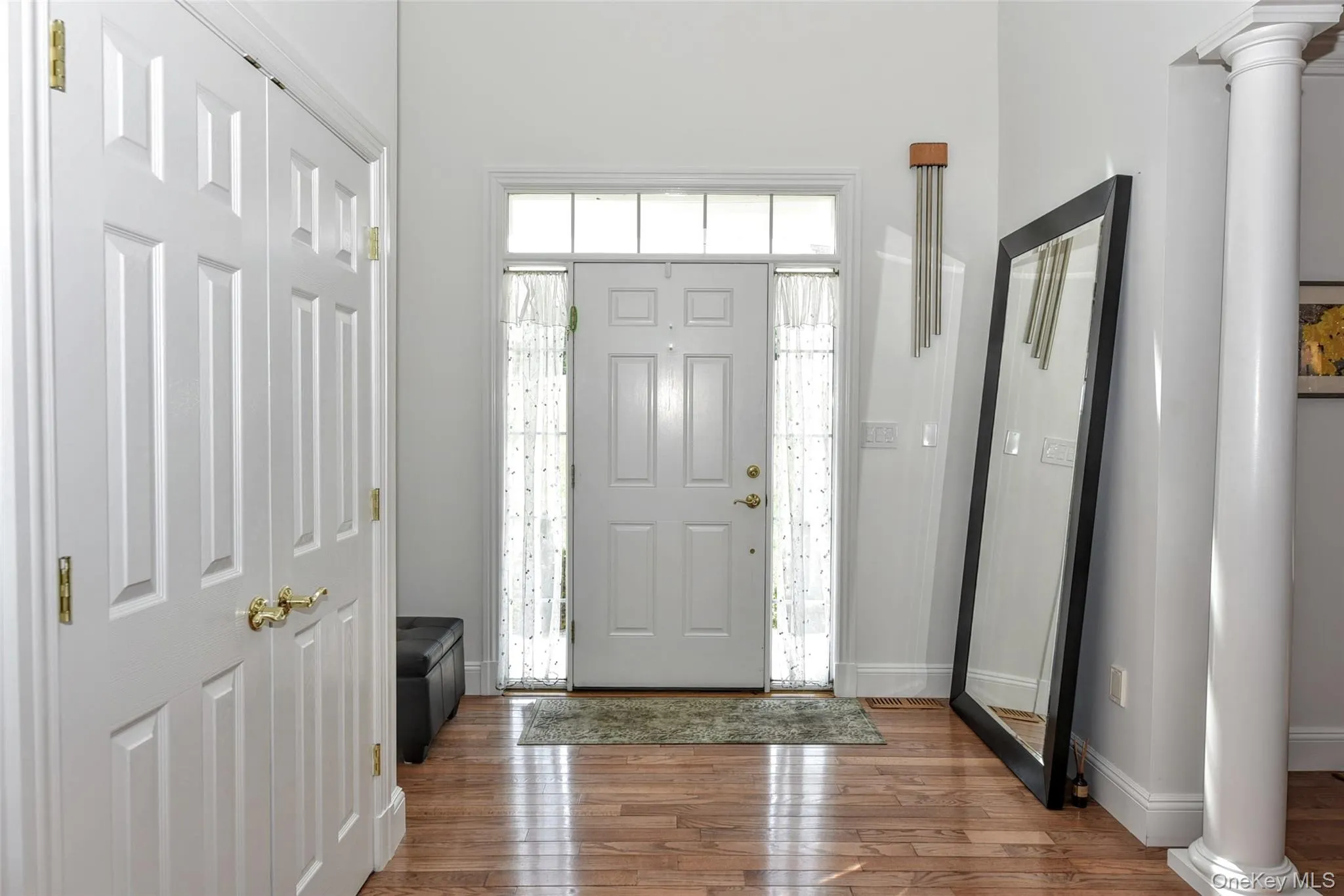 Foyer featuring wood-finish flooring, a white paneled entry door with transom and sidelights, a fluted column, and white bi-fold closet doors with brass hardware Foyer featuring wood-finish flooring, a white paneled entry door with transom and sidelights, a fluted column, and white bi-fold closet doors with brass hardware